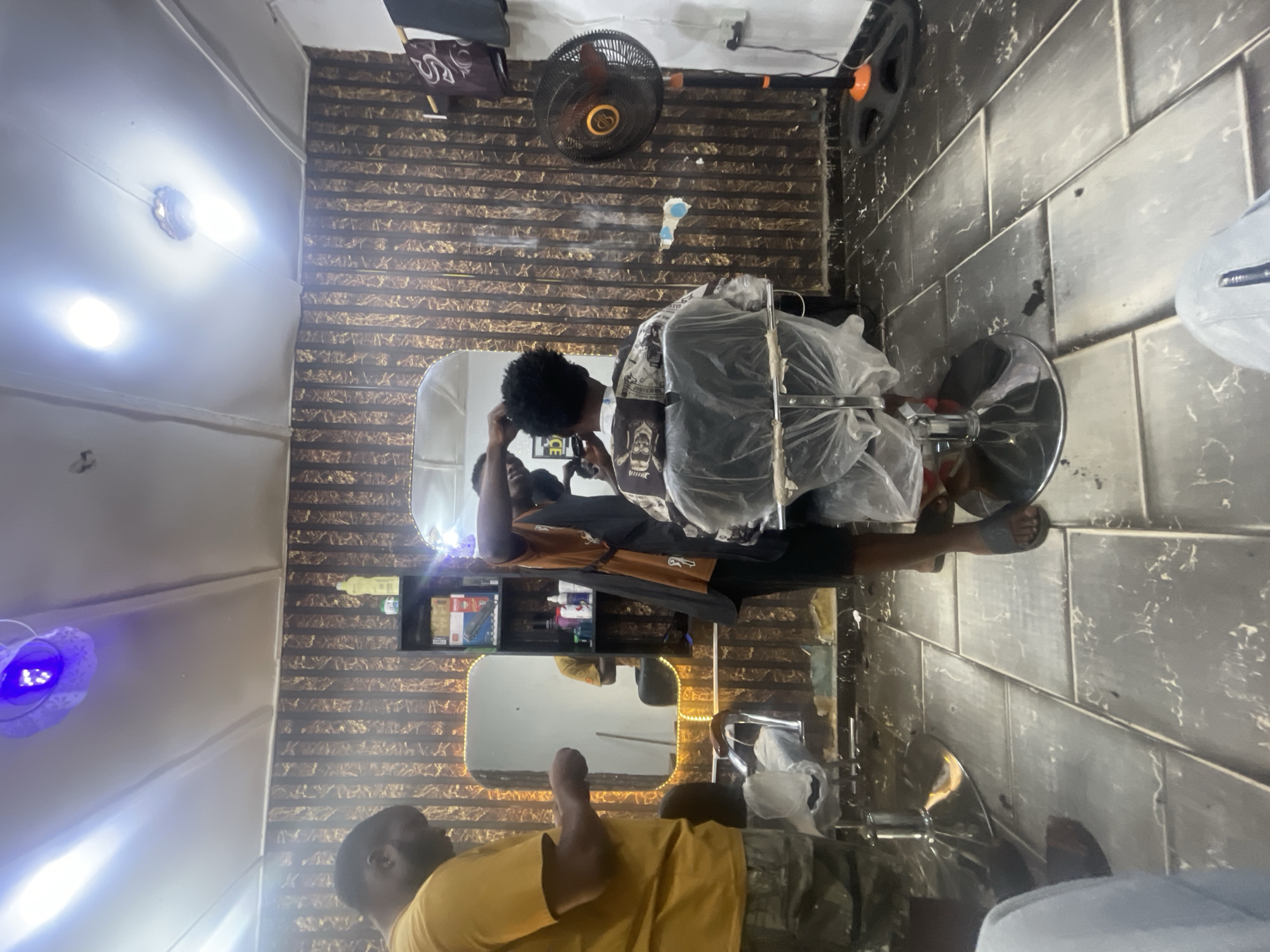 The image depicts the interior of a barber shop, likely in Lokoja, Nigeria, although the location cannot be definitively confirmed. A barber is attending to a customer seated in a barber chair covered with a transparent plastic sheet. The barber is wearing a dark t-shirt. The customer is wearing a brown shirt and black pants. Both are dark-skinned. A mirror is positioned behind the barber chair, reflecting the scene. The shop's walls are decorated with a patterned design. The floor is tiled. Various barbering tools and products are visible on a shelf. A floor fan and a rotating ceiling fan are also present. Another man in a yellow shirt can be seen in the mirror to the left, giving a thumbs up. The lighting in the shop appears to be bright.