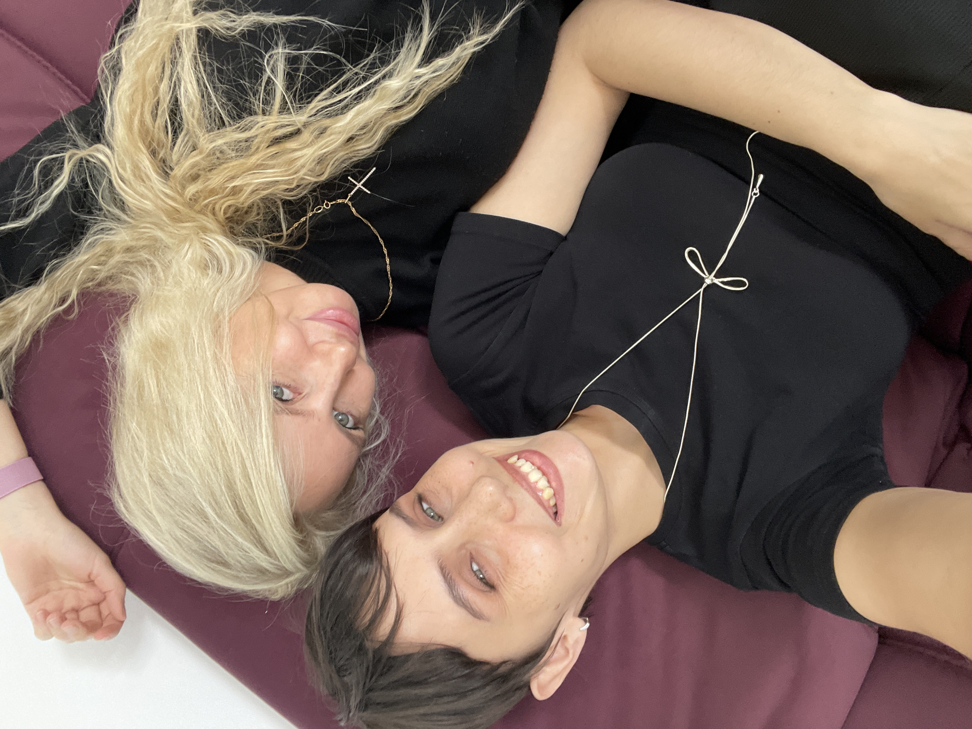 Two women are lying on a purple surface. The woman on the left has long blonde hair and wears a black top with a cross necklace. She has blue eyes and a light complexion. The woman on the right has short dark hair, freckles, and is smiling, revealing her teeth. She is also wearing a black top and a delicate necklace with a bow detail. The image appears to be a selfie taken at an angle. There is no visual evidence to suggest the location is Izhevsk, Russia.
