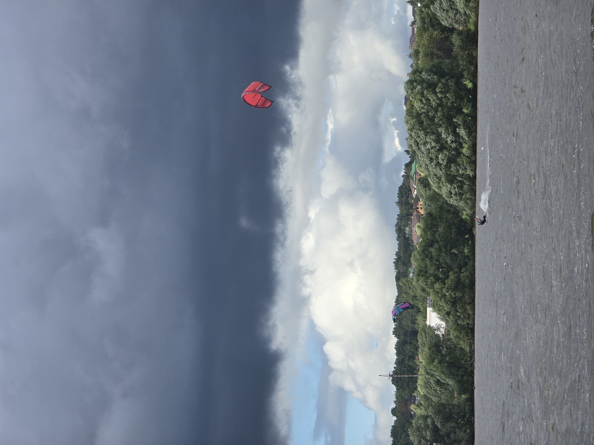 This image depicts a wide shot of a lake with a dark, stormy sky overhead. In the foreground, the rippling water of the lake takes up the lower third of the frame. The middle ground is occupied by a dense line of green trees and vegetation along the shoreline, with houses visible peeking through the foliage. Several power lines also run horizontally across this section.
The sky is dramatic, with large, dark storm clouds dominating the upper half of the image. Patches of lighter, white clouds and a sliver of blue sky are visible between the darker masses.
There are multiple kitesurfers in the water. On the right side of the image, a person is kitesurfing, leaving a spray of water behind them. In the distance, towards the left, another kite surfer is visible. Two large kites are also prominent in the sky: a bright red kite is positioned in the upper right quadrant, and a smaller, multi-colored kite (purple and blue) is further to the left and lower in the frame, over the treeline. The scene appears to be outdoors in a natural setting, likely a recreational area, with the city of Novoseltsevo, Russia suggested by the location.