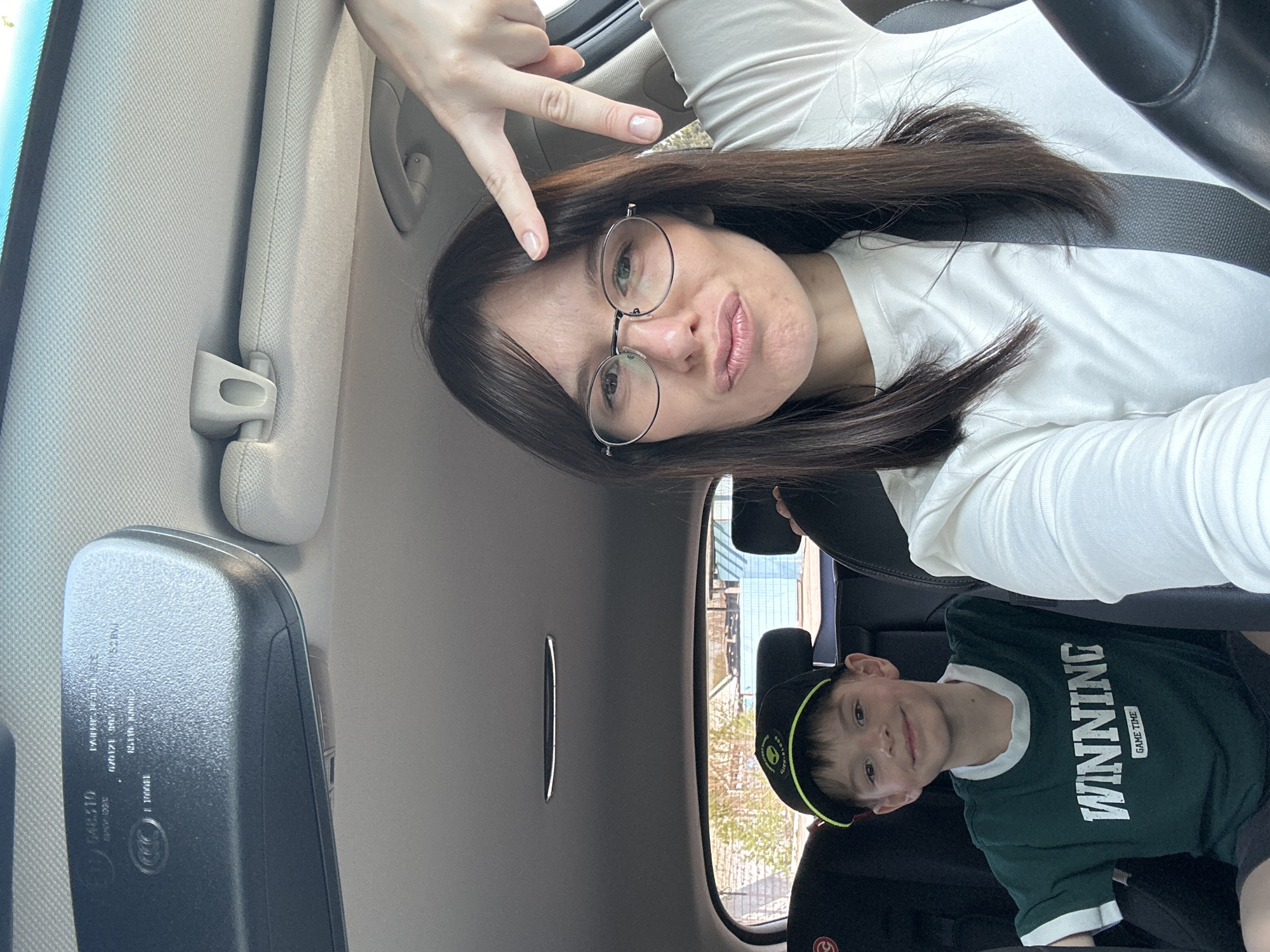A woman and a boy are in a car. The woman, wearing glasses and a white long-sleeved shirt, is in the driver's seat, wearing a seatbelt, and making a hand gesture. The boy, in the back seat, wears a dark green t-shirt that says "Winning" and a baseball cap. The car's interior is visible, including the rearview mirror and sun visor. The exact location within Chaikovsky, Russia, cannot be determined from this image.