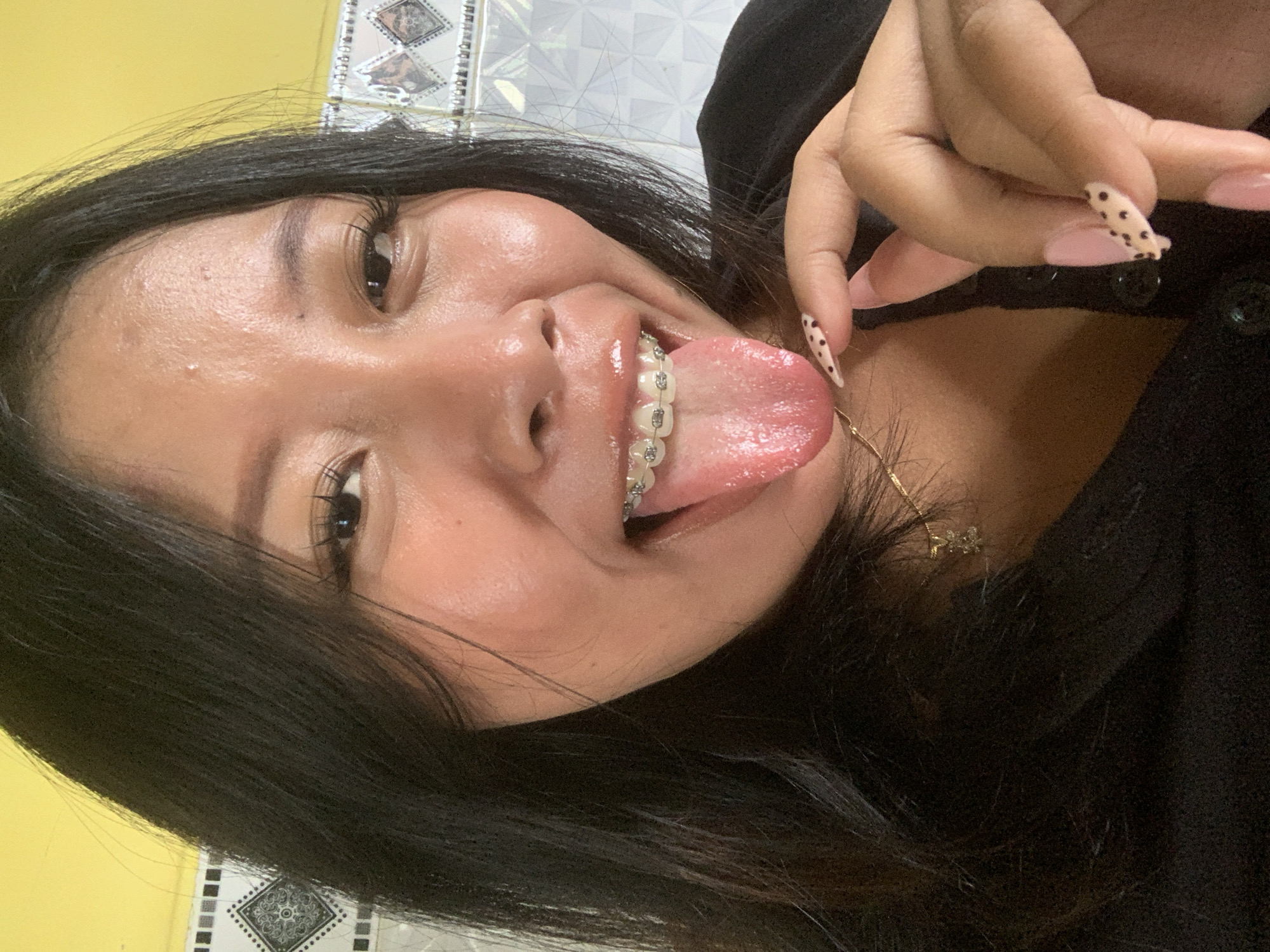 In a close-up selfie, a young Indonesian woman with black hair and braces playfully sticks out her tongue. Her eyes are dark and her lashes are long and accentuated. She wears a black top and a delicate gold necklace. Her fingernails are painted a pale pink with black polka dots. The background shows a yellow wall and patterned tiles. The image appears to have been taken indoors, possibly in a home environment in Jajag, Indonesia, given the distinctive yellow wall and tiled background often seen in residential settings in the region.