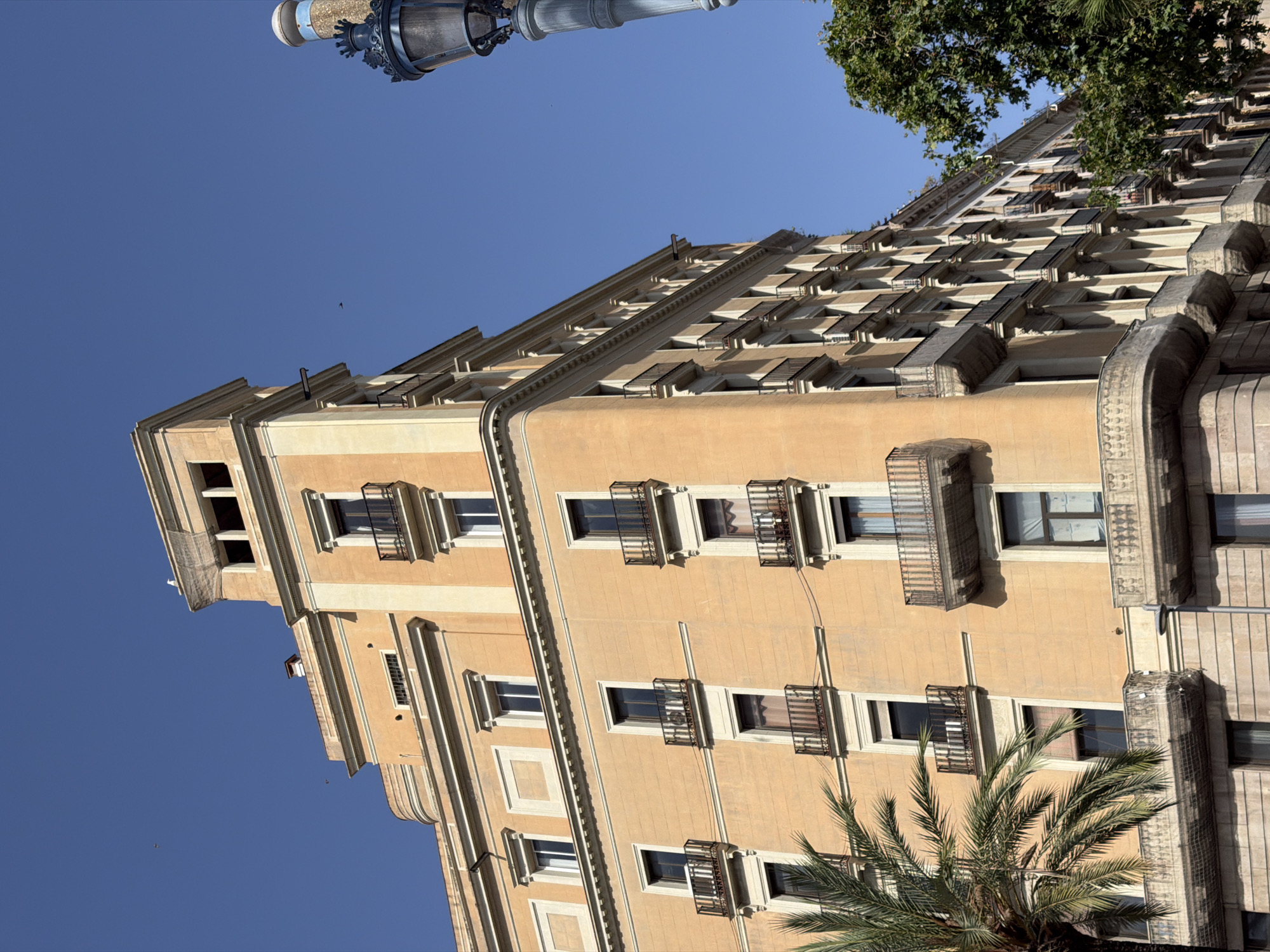 The image captures a detailed view of a tall, peach-colored building in Barcelona, Spain, under a clear blue sky. The building exhibits classic architectural features, including multiple floors with numerous windows, many of which are adorned with black wrought-iron balconies. Some balconies have decorative railings, and others are covered with netting, possibly for protection.
The building's facade is segmented by horizontal cornices and vertical pilasters, creating a sense of order and grandeur. The upper portion of the building culminates in a more elaborate structure with a tower-like element featuring open rectangular recesses.
In the foreground on the left, the fronds of a lush green palm tree are visible, adding a touch of tropical greenery to the urban landscape. To the right, a decorative vintage street lamp with a blue element atop it stands tall. The background is dominated by the deep blue sky, with a few faint specks that could be birds or distant aircraft. The lighting suggests a sunny day, casting subtle shadows on the building's surfaces. The scene is devoid of people, emphasizing the architectural elements and the serene urban environment.