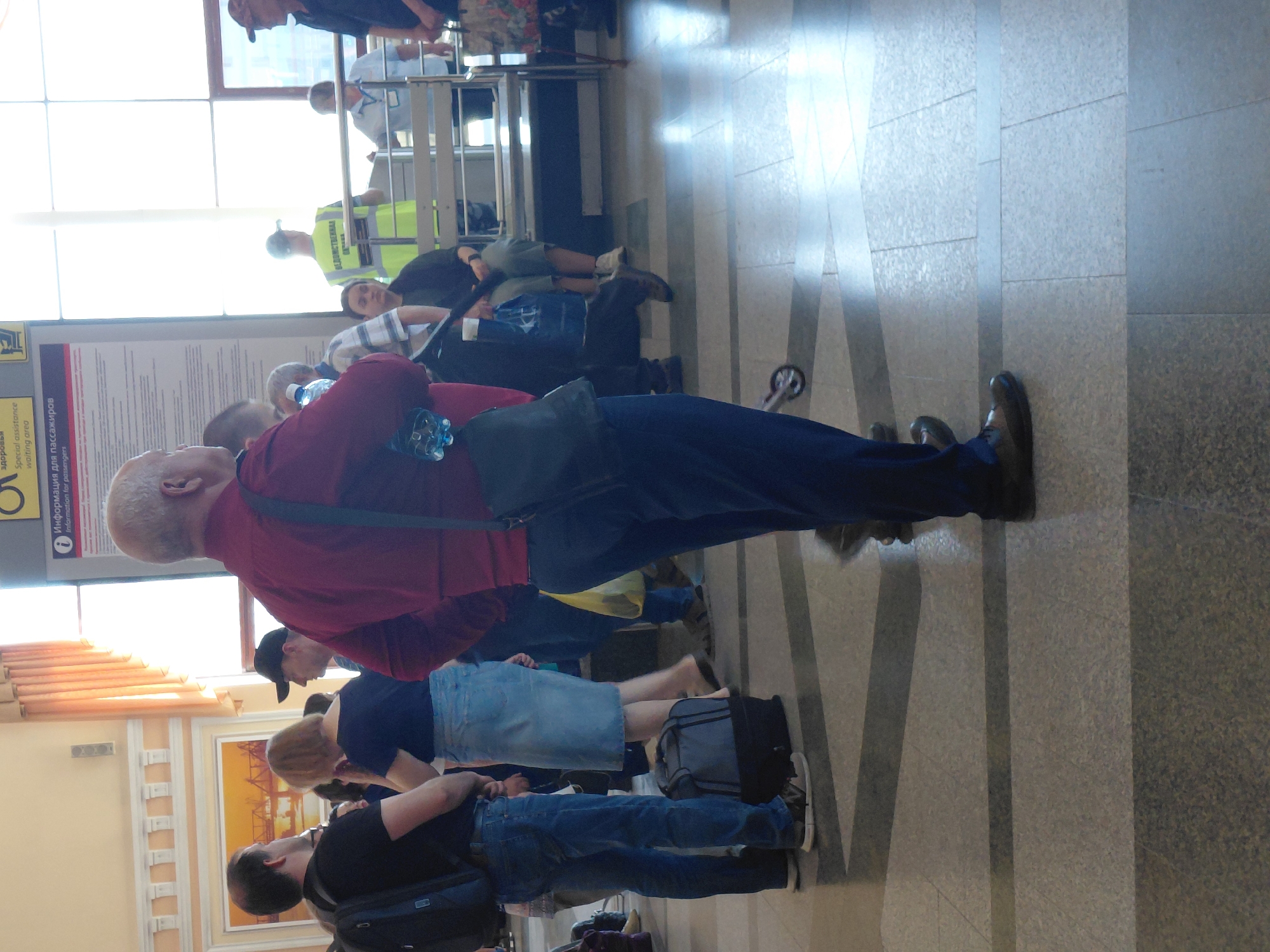 A man in a burgundy shirt and dark pants stands facing right, with a dark bag slung over his shoulder. He is holding a water bottle in his right hand. To his left, a woman in a blue dress and light-colored skirt stands facing left, with a gray suitcase at her feet. To the left of the woman, a man in a black t-shirt and blue jeans stands facing left, with a black backpack on his back. In the background, several other people are visible, some sitting on benches and others standing. There is a sign with Cyrillic text and an image of a circle with a diagonal line through it. The floor is tiled with gray and light gray squares. The environment appears to be an indoor public space, possibly a train station or airport waiting area.