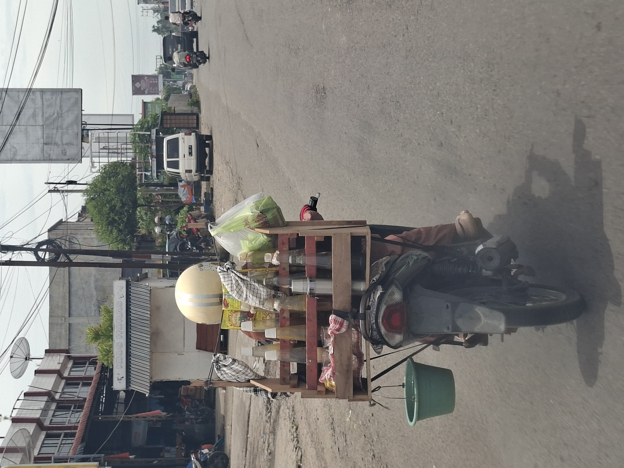 A roadside scene in Meukokuthang, Indonesia, shows a motorcycle laden with a wooden rack holding bottles, some draped with cloths. The motorcycle also carries a helmet, a plastic bag of produce, and a green bucket. Buildings and other vehicles are visible in the background.