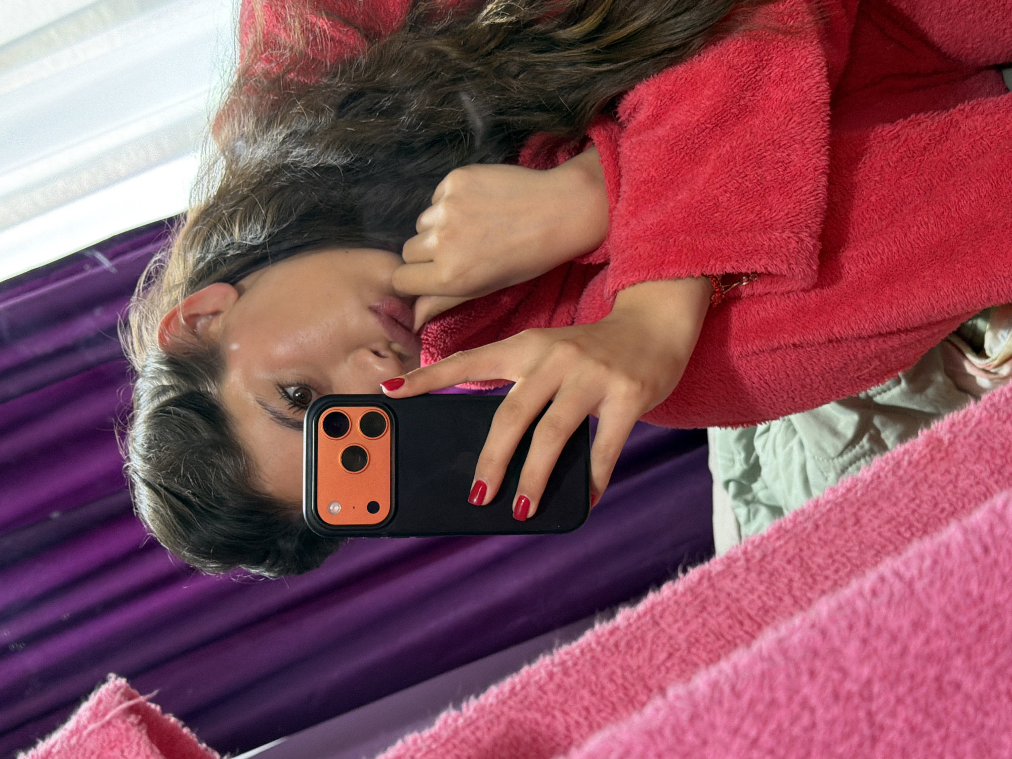 A woman in a red fuzzy robe is taking a selfie in a mirror. She is holding a black phone with a light orange square around the camera lenses. The reflection shows a purple curtain and pink fuzzy material. The image does not provide any information relating to the city of Dragichevo, Bulgaria.