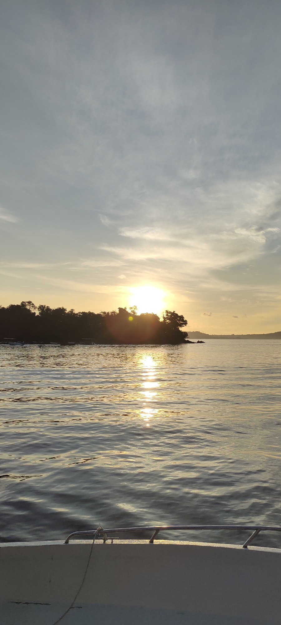 The image captures a tranquil sunset scene over the ocean, likely near Sabang, Indonesia. The sun, a bright orb, is positioned low on the horizon, partially obscured by a silhouetted island covered in dense vegetation. The sky above is a gradient of soft blues and greys, with wispy clouds catching the warm, golden light of the setting sun. A shimmering reflection of the sun dances on the rippling surface of the water, creating a path of light that stretches towards the viewer. In the foreground, the edge of a boat's railing is visible, suggesting the image was taken from a vessel on the water. The overall atmosphere is one of peace and natural beauty, characteristic of coastal landscapes in tropical regions.