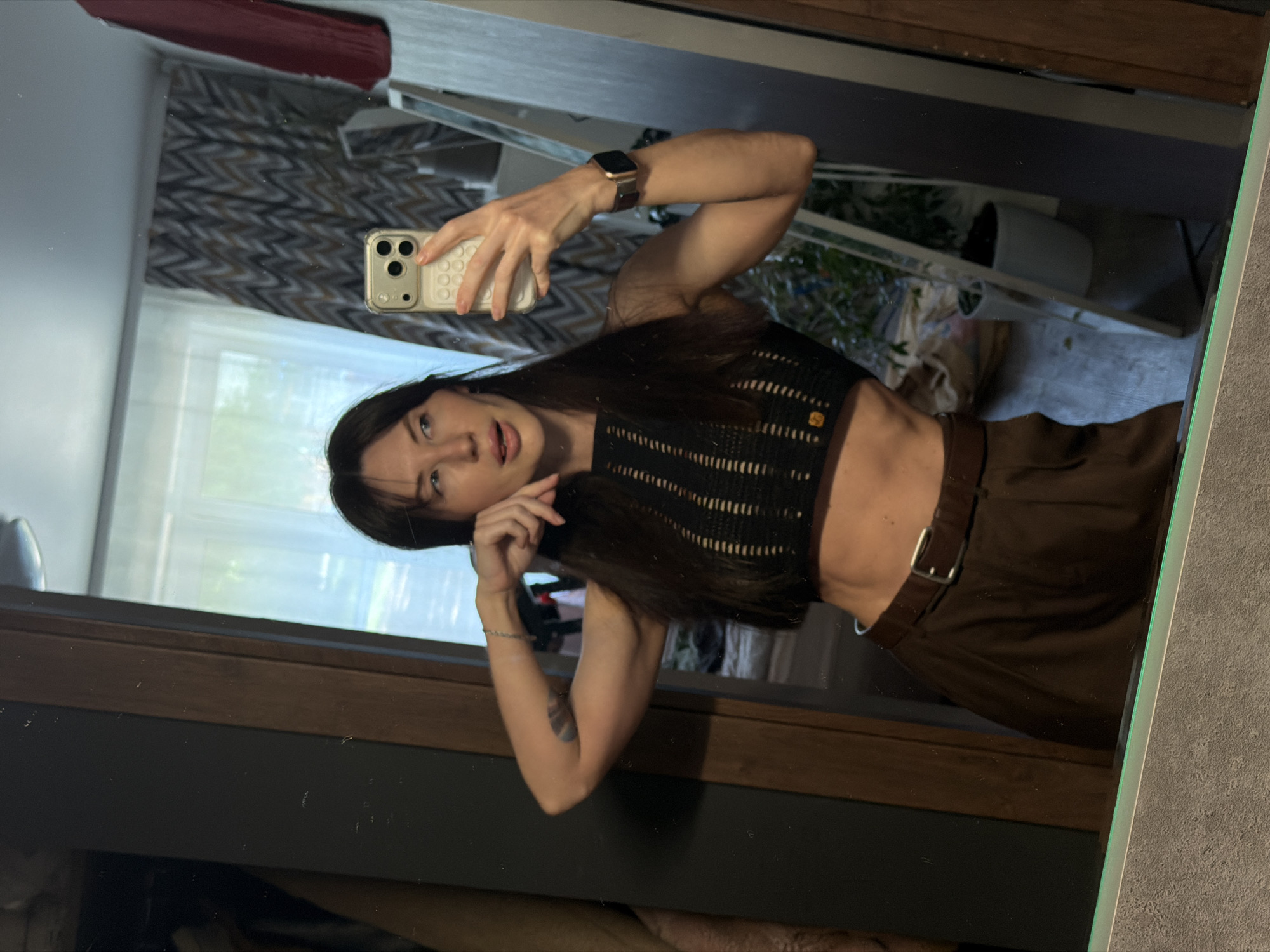 A woman is reflected in a mirror. She holds a cell phone up to take a selfie, and she is wearing a cropped black top, and brown trousers with a belt. A brown curtain is visible. A window is visible. The mirror is against a wall. The woman wears an Apple watch. The image was taken in Perm, Russia.