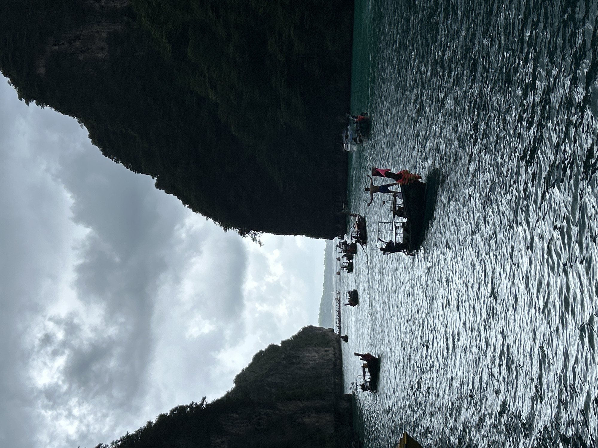 The image depicts a dramatic scene in Haad Yao, Thailand, under a heavily overcast sky. The water is a turbulent, deep teal, reflecting the muted light. Towering, dark green limestone karsts flank a narrow channel, their sheer faces covered in dense vegetation. Several traditional Thai longtail boats are scattered across the water. In the foreground, a man stands with arms outstretched on the bow of a boat, facing away from the viewer, with a pink flag visible on the boat. Other boats, some with passengers, navigate the channel, their silhouettes stark against the water. In the distance, more boats and a hazy coastline suggest a populated area. The overall atmosphere is one of awe-inspiring natural beauty tempered by a sense of impending weather.