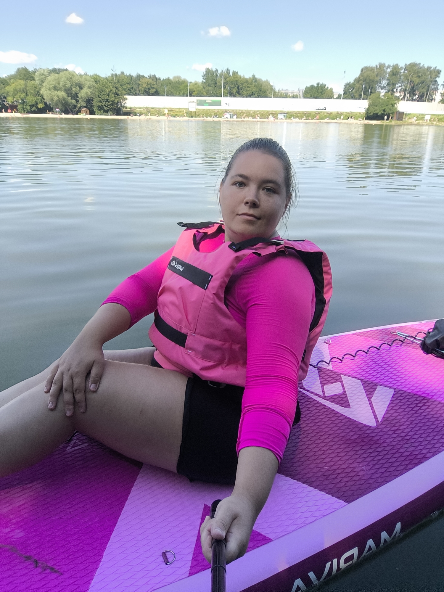 A woman in a pink life vest and pink long-sleeved shirt sits on a pink paddleboard on a lake. She is holding a selfie stick. In the background, there is a sandy beach with trees and buildings. The sky is blue with some clouds. The image is taken on a sunny day. The paddleboard is labeled "AVIGAM".