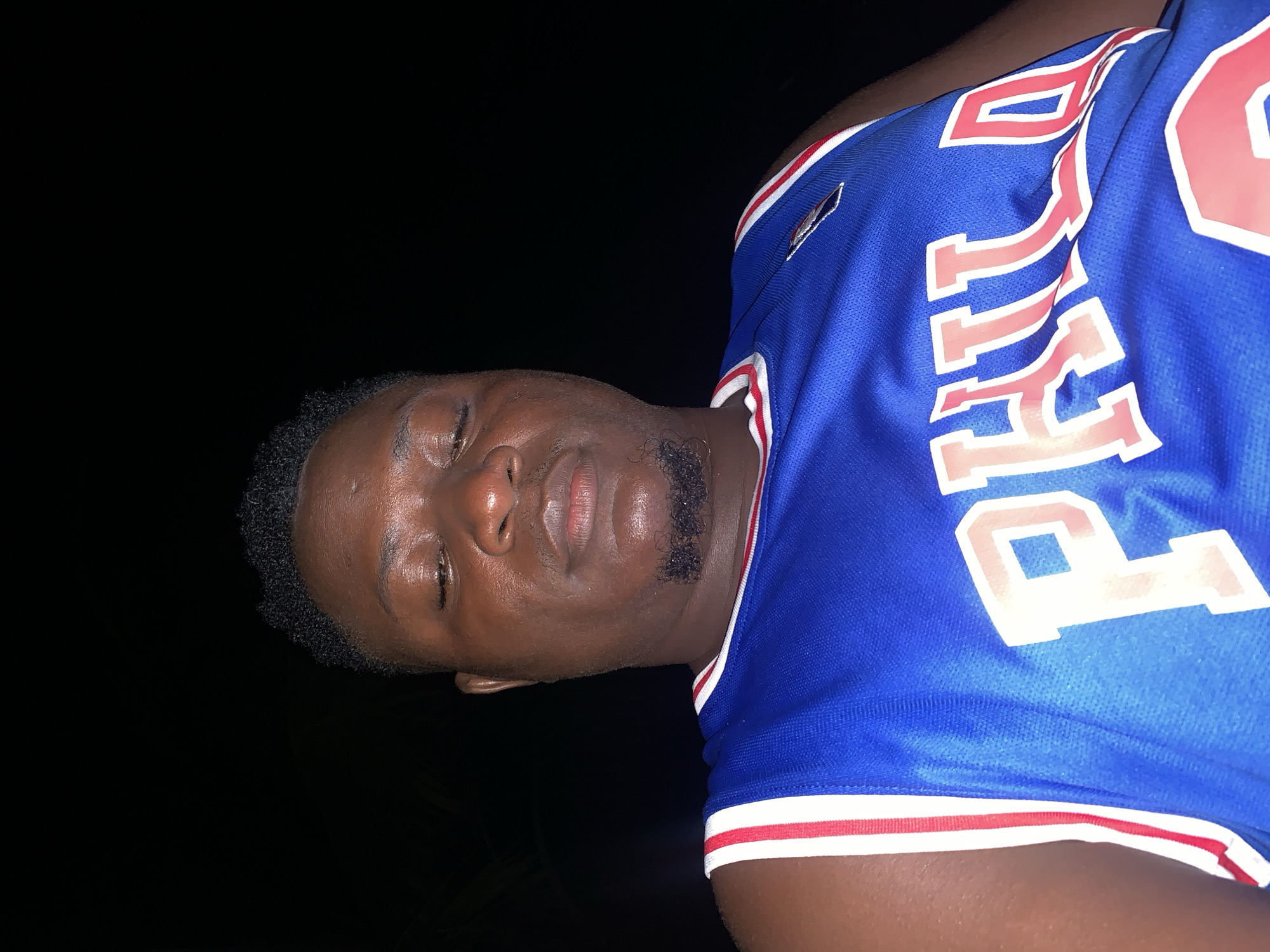 A close-up, low-angle selfie shows a Black man in a blue basketball jersey with red and white trim. The jersey has the word "PHIL" printed in large, red, white-outlined block letters across the chest. The man's eyes are closed, and he has a light beard and goatee. The background is dark and out of focus, suggesting the photo was taken at night. The scene appears to be outdoors, possibly in Akwanga, Nigeria, as indicated by the provided context.
