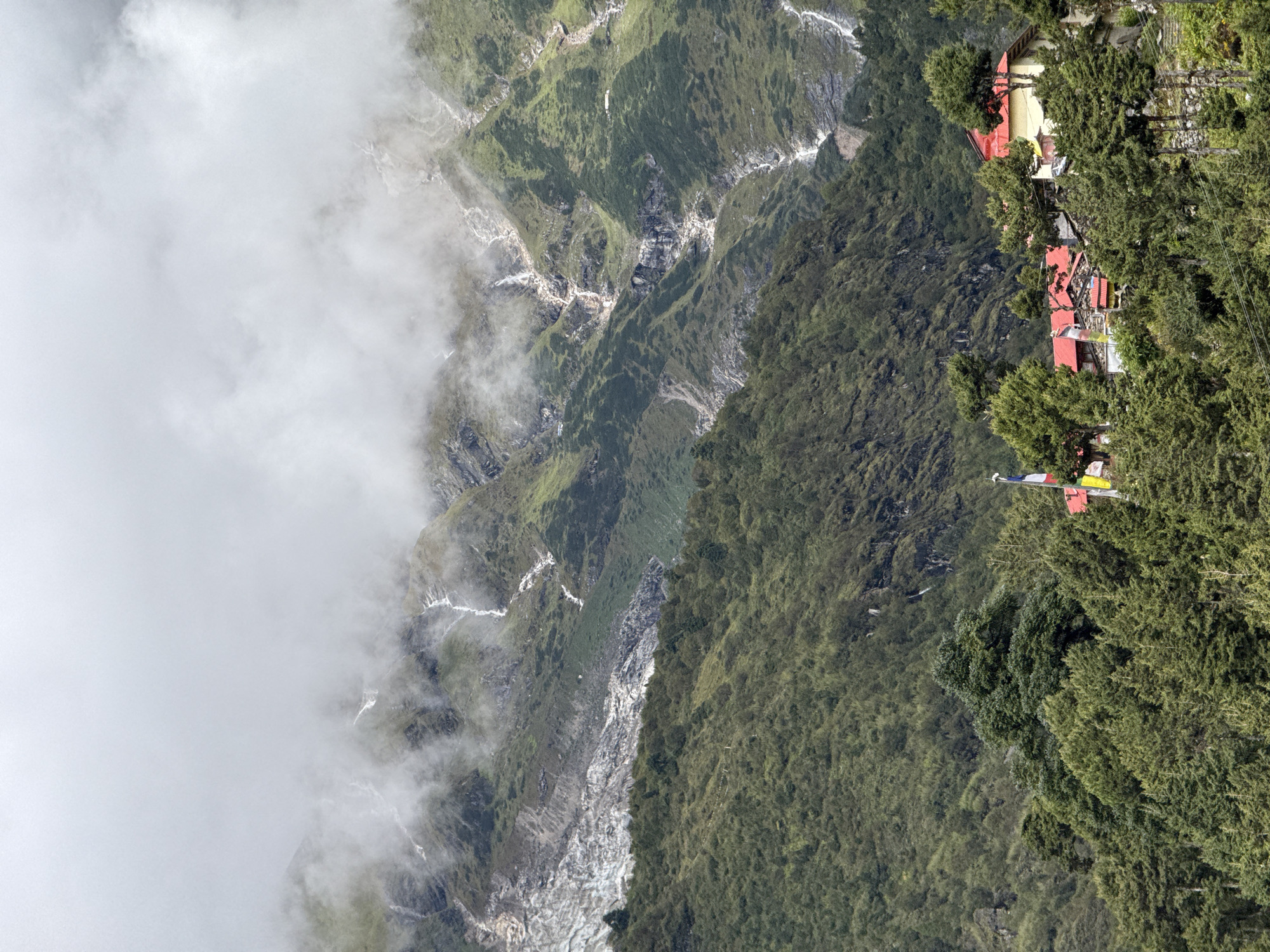 The image shows a view of the village of Samagaon, Nepal. In the foreground, several buildings with red roofs are nestled amongst lush green trees. A few prayer flags are visible, adding color to the scene. In the background, a majestic mountain range rises, with some areas covered in mist. White streaks, likely waterfalls or snowmelt, cascade down the rocky slopes. A glacier can be seen on the upper left side of the mountain. The overall atmosphere is one of natural beauty and tranquility.