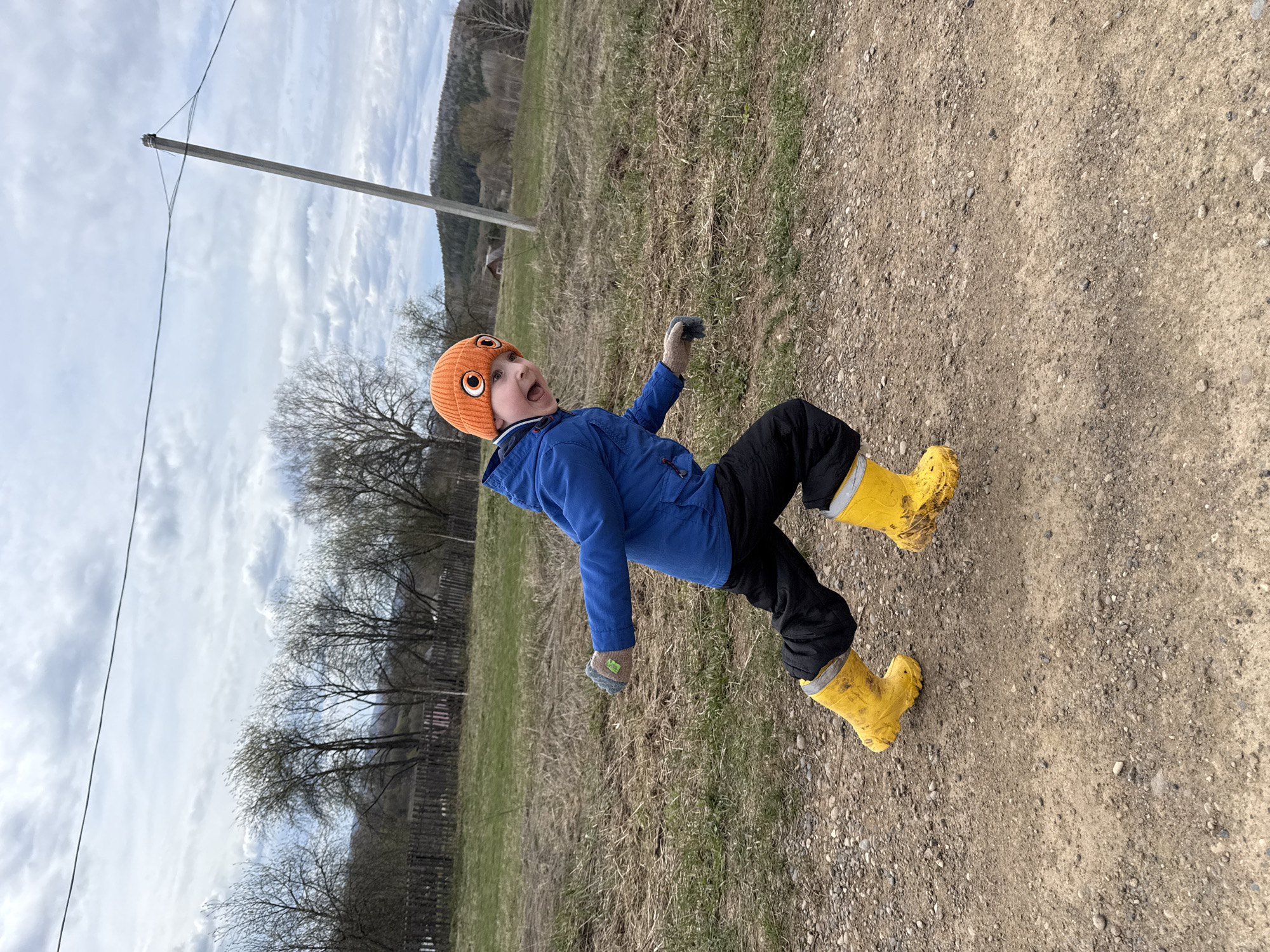 A child wearing an orange hat, blue jacket, black pants, and yellow boots is on a dirt path in what appears to be a rural area of Усть-Тунтор, Russia. The child is wearing gloves. The sky is cloudy, and there are trees, grass, and a telephone pole visible in the background.