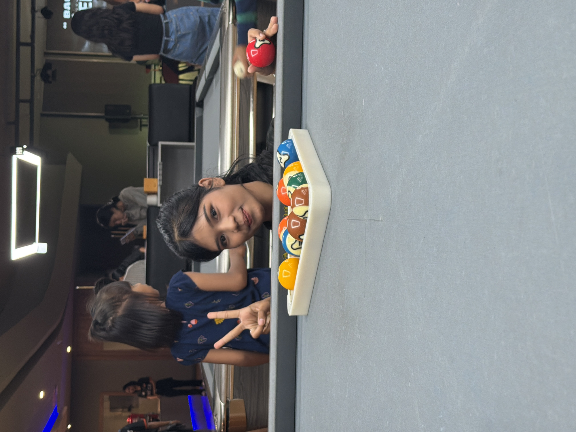 The image depicts a dimly lit indoor environment, likely a pool hall, in Kuta Selatan, Indonesia. A young woman with dark hair pulled back in a ponytail is in the foreground, leaning over a pool table and looking at the camera with a slight smile. She is wearing a black top. To her left, a young girl with dark hair in a similar hairstyle is standing, also leaning over the pool table and holding up two fingers in a peace sign. She is wearing a dark blue t-shirt with a pattern.
On the pool table, a white rack holds several billiard balls, including orange, brown, and blue balls with visible numbers. To the right of the frame, another hand is visible, holding a red billiard ball with the number 7.
In the background, other individuals are present, though less clear. One person is seated at a table in the middle ground. Further in the background, another person is standing, wearing a black t-shirt and denim shorts, and another person is visible from the back wearing a dark top and denim jeans. The environment appears to be a leisure space with a pool table as the central element.