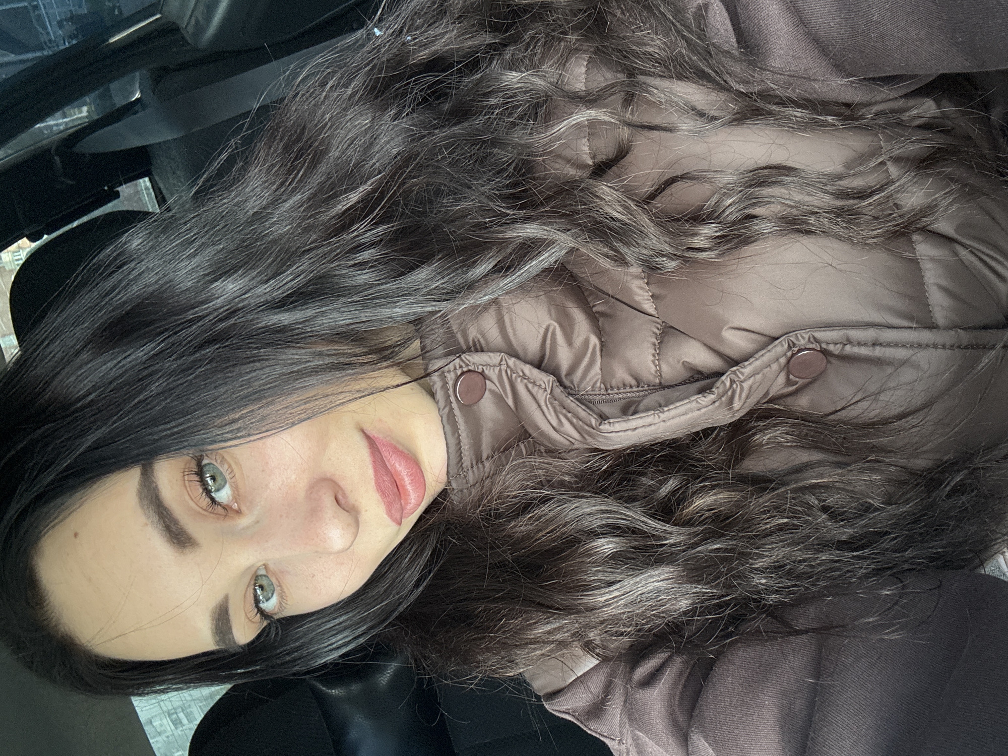 The image presents a woman with long, wavy, dark brown hair and light-colored eyes. She is wearing a brown puffer jacket with visible buttons. The background suggests she is inside a vehicle, possibly a car, with glimpses of the exterior environment visible through the window. The location is not definitively identified as Chaikovsky, Russia, from the visual information provided. There are no discernible actions or interactions occurring in the frame. The environment is generally overcast, as suggested by the lighting in the vehicle and visible outside.