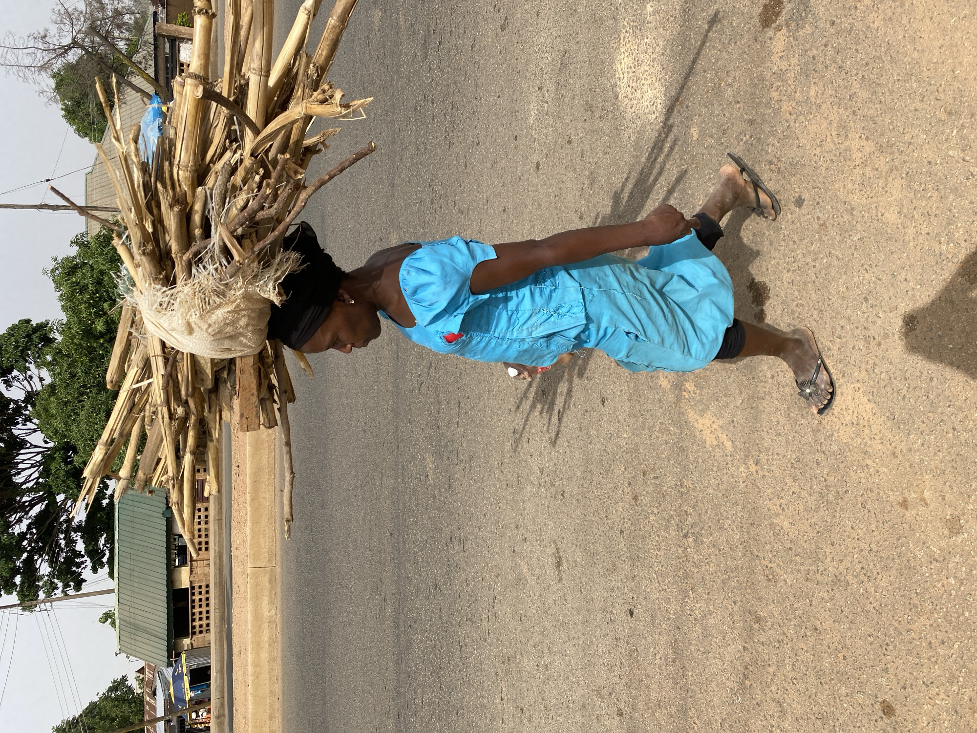 A woman walks along a paved road in Bukuru, Nigeria, carrying a large bundle of dry stalks and branches on her head, secured with a cloth or bag. She wears a light blue, short-sleeved dress and dark footwear; her legs are visible. The road is wide, and a building with a green roof is seen to the left of the frame, along with some trees and power lines in the background. The lighting suggests it is daytime.