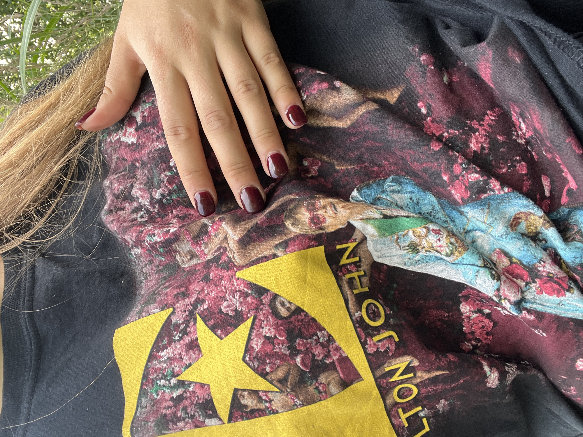 In an outdoor setting with blurred greenery in the background, a person's torso is visible, wearing a black t-shirt with a colorful graphic print. The graphic features a large yellow star with an outline, partially obscuring a portrait of Elton John. Text within the star reads "E", and below it, "ELTON JOHN" is printed in yellow. The t-shirt is adorned with a floral pattern in shades of pink and red, and Elton John is depicted wearing a blue jacket with floral embellishments and pink sunglasses. The person's right hand is resting on the t-shirt, with fingernails painted a dark red color. The image appears to be taken from a medium shot, focusing on the upper body and the t-shirt design. The specific location within Дзямідавіцкі сельскі Савет, Belarus cannot be determined from the visual information provided.
