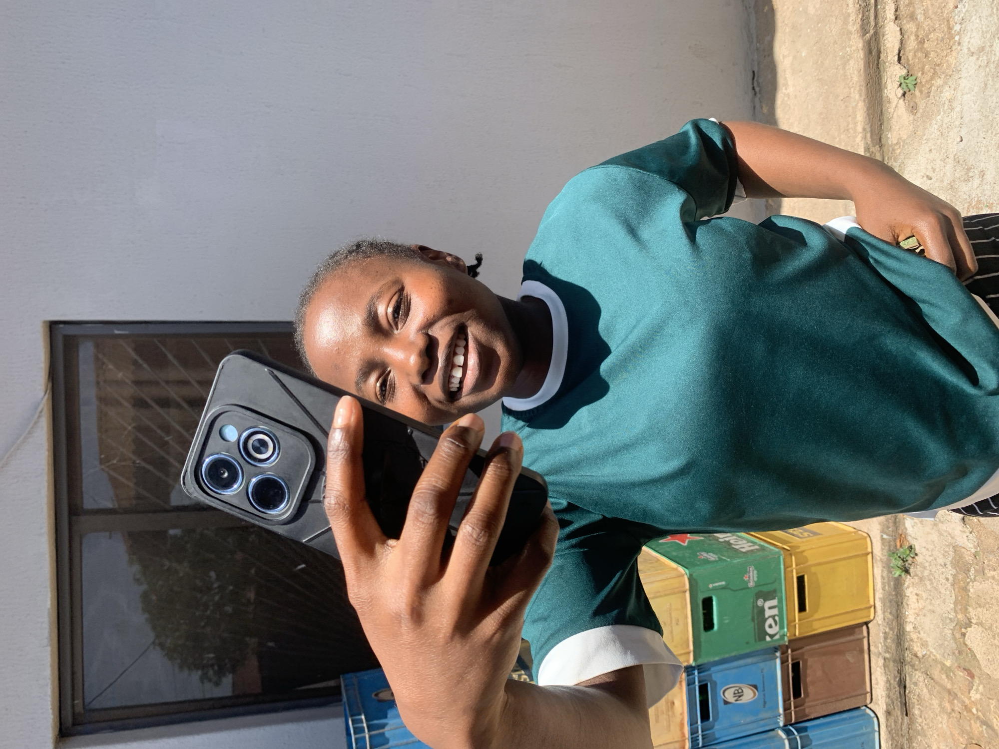 A person is captured in a brightly lit outdoor selfie, holding a modern smartphone with multiple visible camera lenses in a dark case. The subject, who appears to be a young individual with dark skin and short dark hair, is smiling broadly with teeth showing, looking directly at the camera with a joyful expression. They are wearing a teal or dark green t-shirt with a white collar and trim.
The background features a light-colored wall, possibly white or light grey, which casts strong shadows from the direct sunlight, suggesting a sunny day, likely midday or early afternoon. A dark-framed window is visible in the wall, reflecting some foliage. Below the window, several plastic crates are stacked on the ground. These crates are of various colors, including green, yellow, and blue. Visible text on the crates includes "Heineken" on a green crate and "NB" on a blue crate. The ground beneath the crates is a rough, textured surface, possibly concrete or packed dirt, with a small patch of green vegetation. The setting appears to be an informal outdoor area, consistent with a location in Jos, Nigeria.