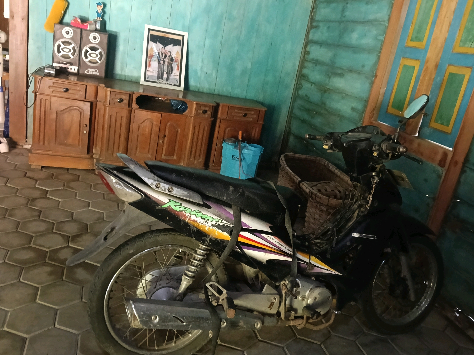 The image shows an indoor scene in what appears to be a residential or storage area in Nglungger, Indonesia. In the foreground, a black motorcycle is parked on a hexagonal tiled floor. The motorcycle has a basket attached to the handlebars and a strap draped over its seat. The rear wheel of the motorcycle is visible, along with its exhaust pipe and part of the engine.
Behind the motorcycle, a wooden cabinet with multiple drawers and doors is positioned against a teal-colored wooden wall. On top of the cabinet are two speakers and a framed photograph of a wedding party. To the right of the cabinet, a blue bucket with a sprayer attached is on the floor. Further to the right, a wooden door with blue and yellow paneled sections is visible. The overall environment appears somewhat cluttered, suggesting a lived-in or functional space.