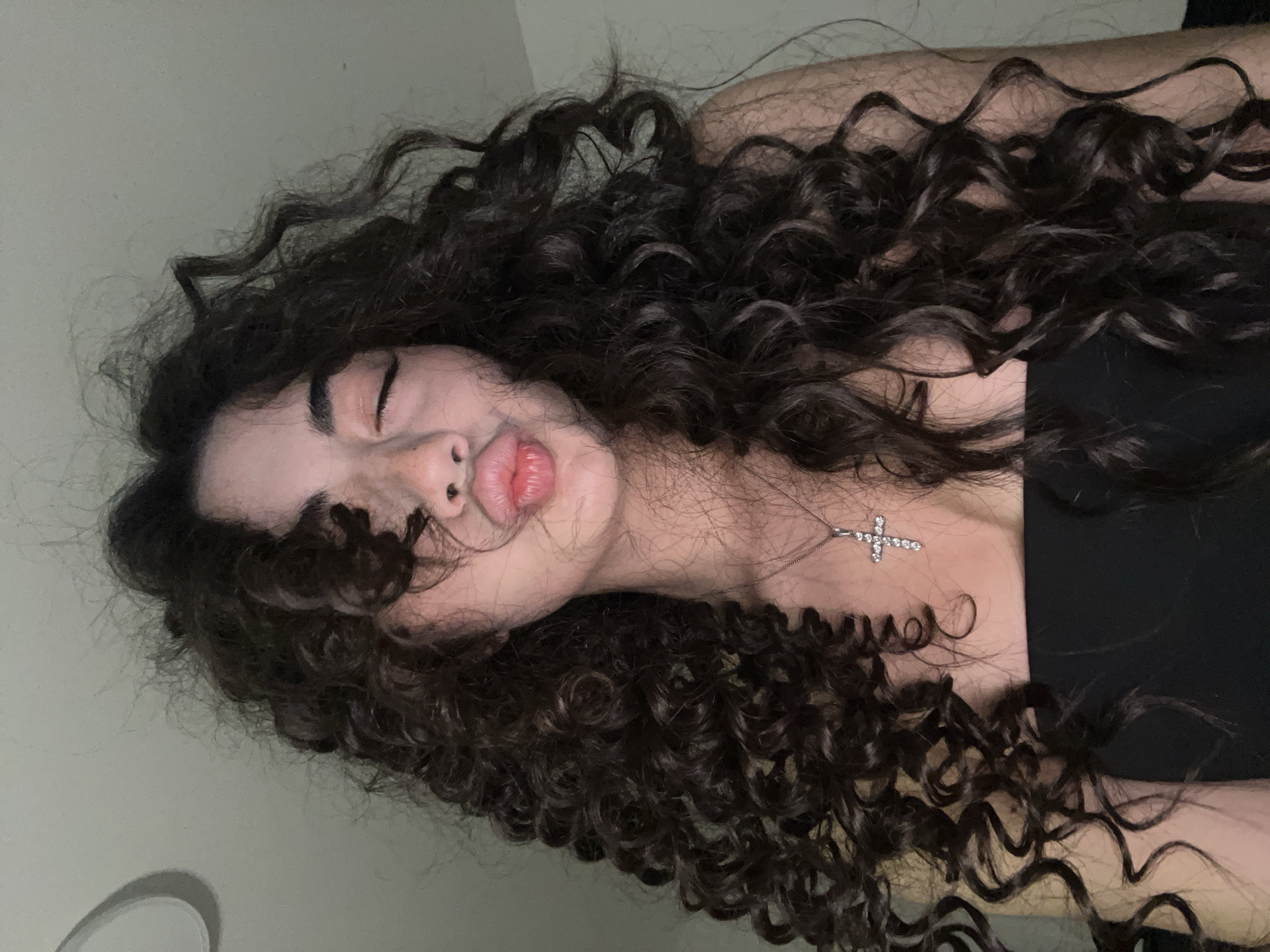 The image shows a close-up of a young woman with dark, curly hair. She is wearing a black strapless top and a silver necklace with a cross pendant. Her eyes are closed, and her lips are pursed as if she is about to kiss. The background is a plain, light-colored wall with a circular light fixture visible on the upper left. There is no indication of the location being in Chotěšov, Czechia. The lighting appears to be artificial and soft.