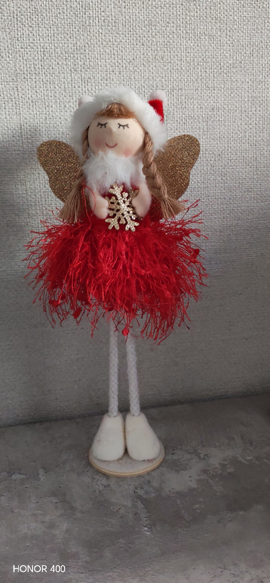 The image shows a handcrafted Christmas angel doll. The doll has a soft, light skin-toned face with closed eyes and a small, smiling mouth. It wears a white faux fur-trimmed hood with a small red Santa hat detail on the side. The doll has golden braided hair and gold glitter wings. It has a white faux fur collar and holds a gold snowflake ornament. The doll is wearing a red, fluffy, fringed skirt. Its legs are white yarn-like material, and its feet are white and rounded. The doll is standing on a round wooden base. The background is a light gray wall. The bottom of the image has "HONOR 400" printed. The image does not provide information that could determine the location in Noviy, Russia.