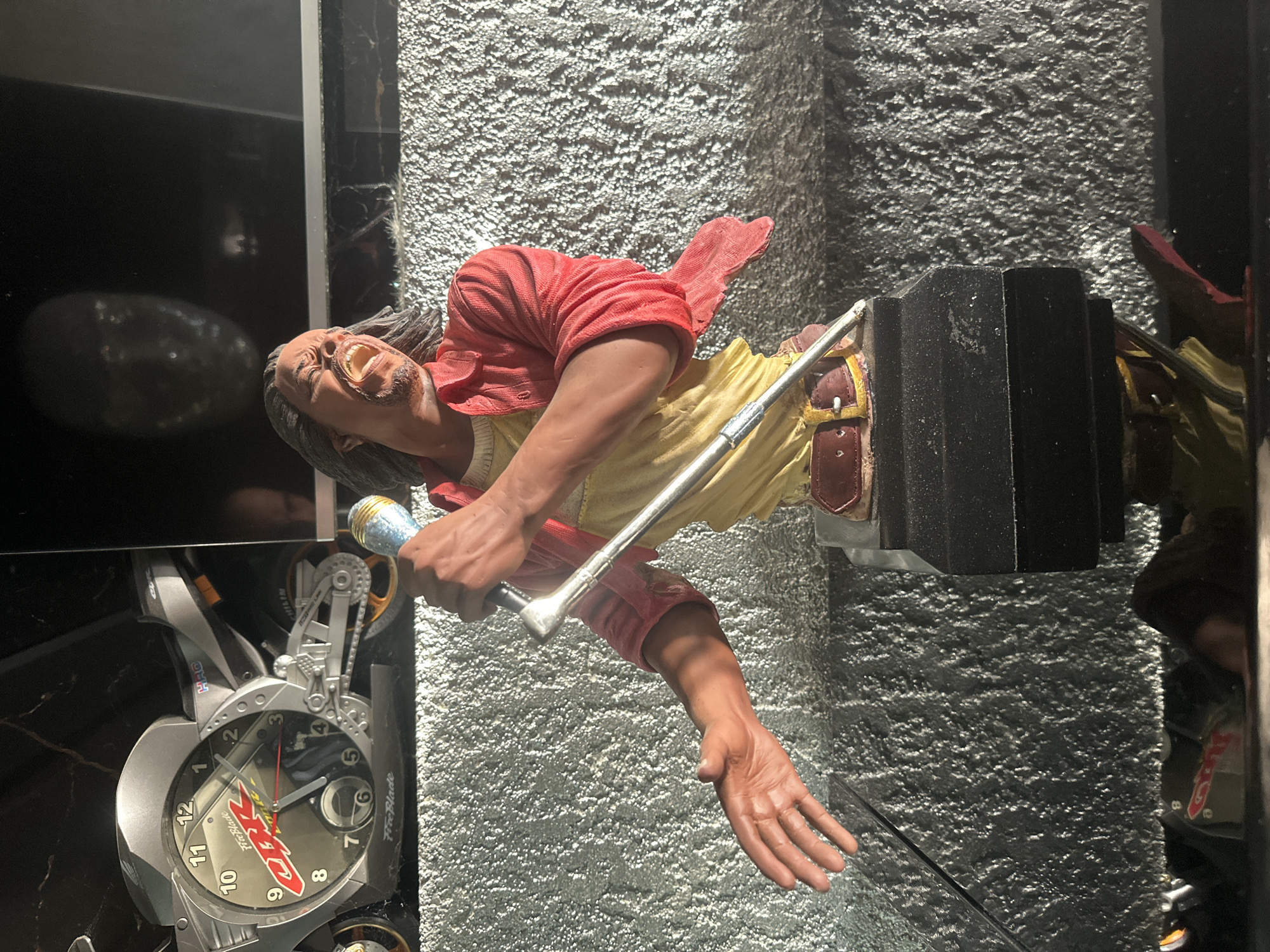 The image shows a close-up view of a detailed figurine of a man singing into a microphone. The figurine is wearing a red open shirt over a yellow garment and a brown belt. He has long dark hair and his mouth is open in a singing expression. The figurine is placed on a black base. In the background, there is a motorcycle-shaped clock, with the brand "CBR" visible. Further back, a blurred reflection of a face is visible on a dark surface. The surface on which the figurine and clock are placed appears to be textured and grey. The setting is indoors, with lighting that creates highlights and shadows. There is no indication of the city of Brca, Montenegro, in this image.