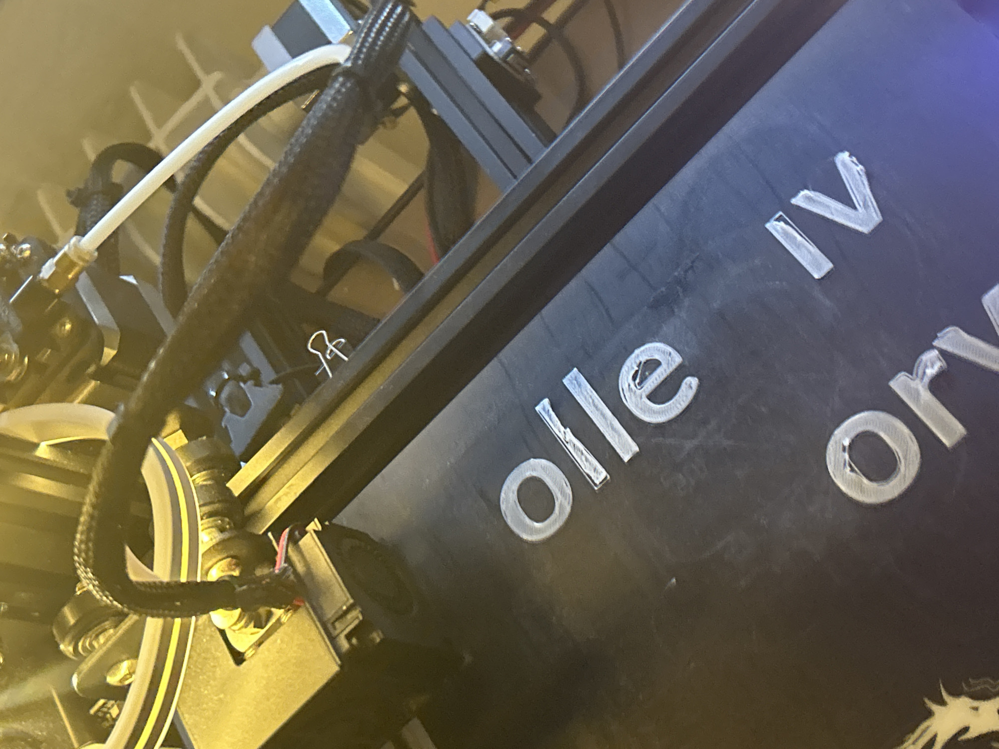 The image shows a portion of a 3D printer, specifically the hot end assembly and part of the frame. The frame has the words "olle IV" printed on it in raised white lettering. A white PTFE tube runs from the extruder to the hot end, and is covered in black wire loom. The printer appears to be in an indoor environment with artificial lighting. This image does not depict Strasbourg, France.