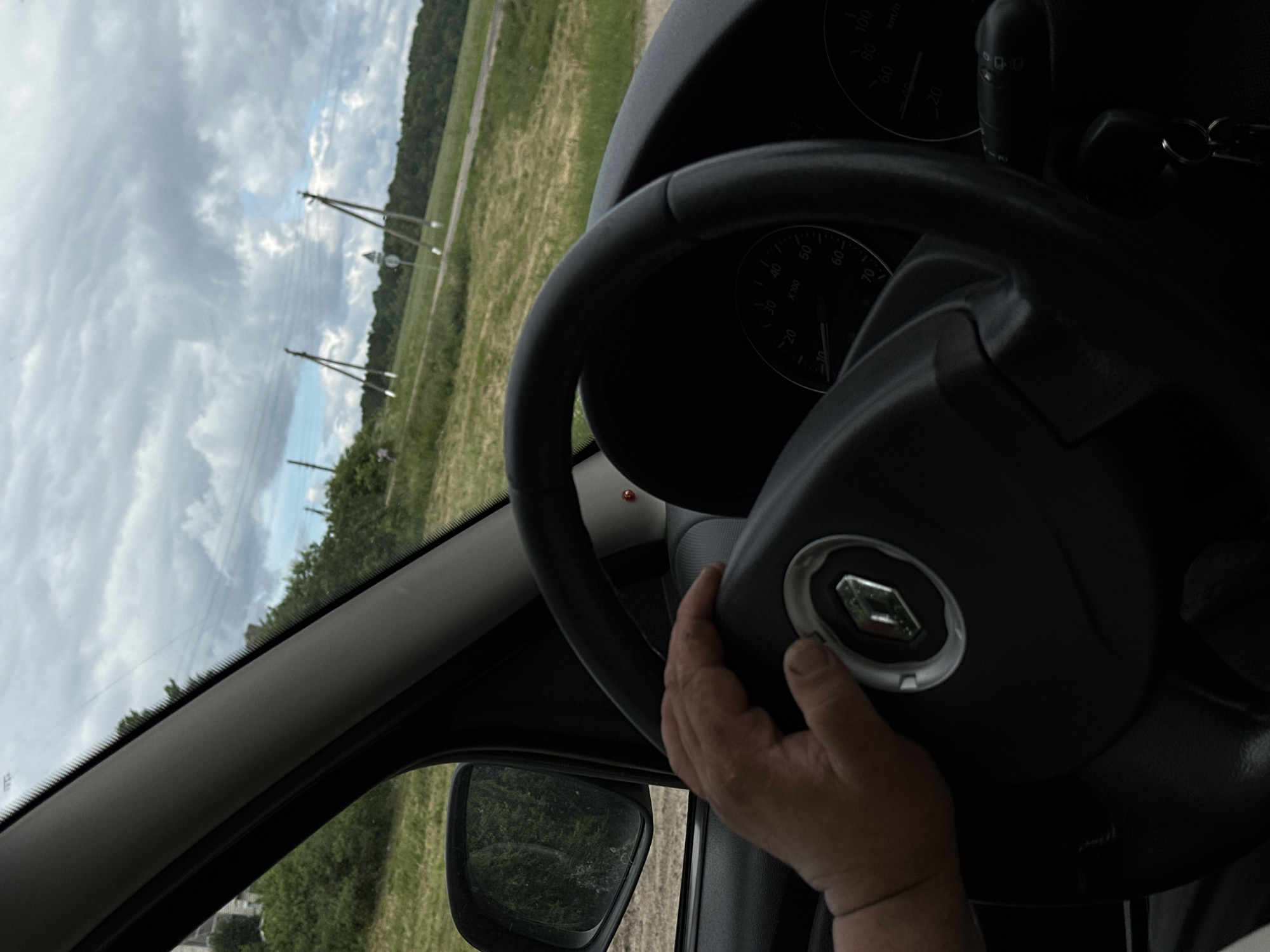 The image shows a first-person perspective of a person driving a car, with their hand on the steering wheel. The steering wheel has a Renault logo in the center. The car's dashboard instruments, including the speedometer, are partially visible. Outside the car, a rural landscape with green fields, trees, and power lines under a cloudy sky can be seen. The location is identified as Ilyanskiy selskiy Sovet, Belarus.