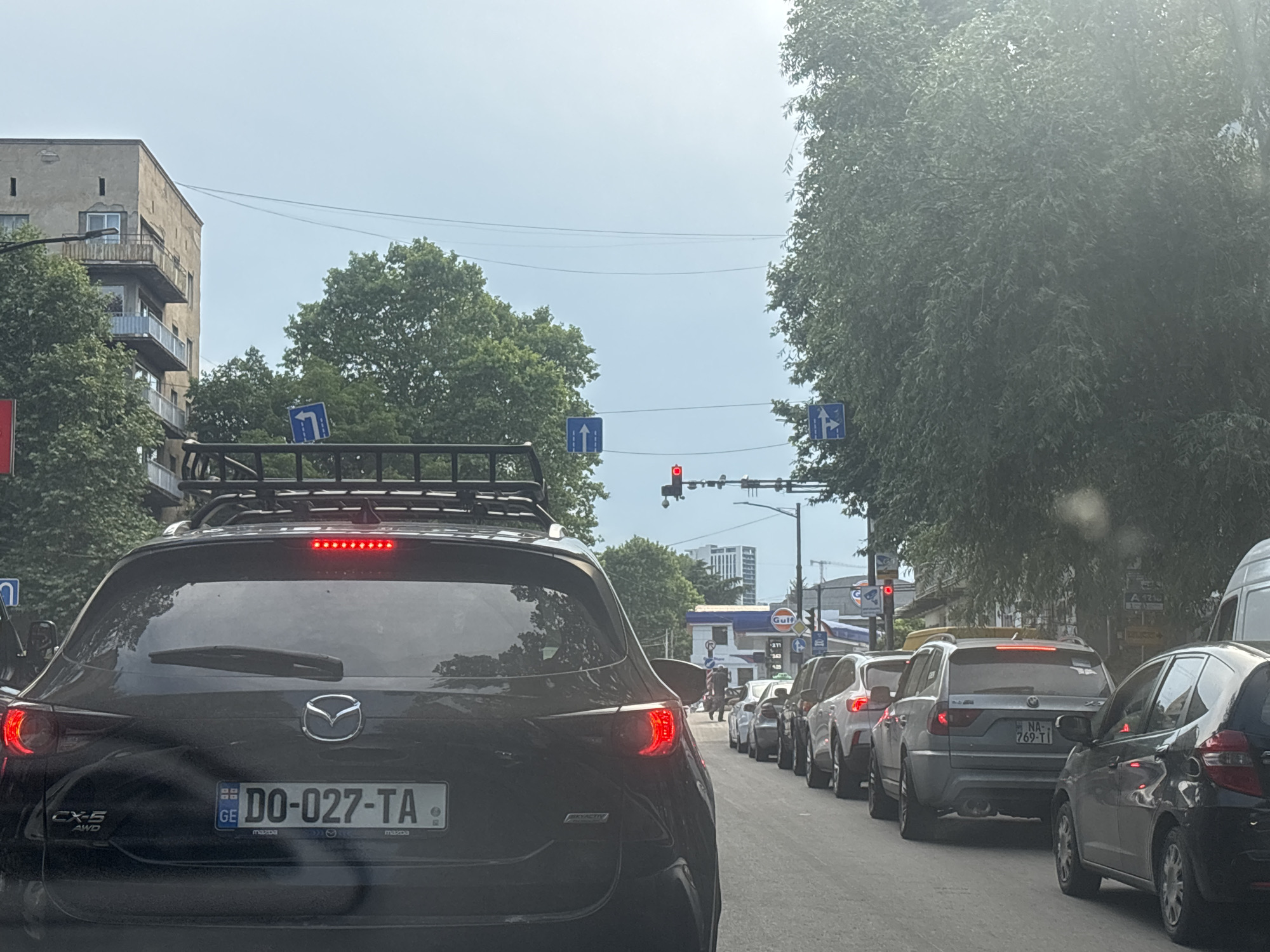 This media file captures a street scene in Tbilisi, Georgia, likely during daylight hours given the bright sky. The foreground is dominated by the rear of a black Mazda CX-5, its taillights illuminated. A roof rack is mounted on top of the vehicle. The license plate of the Mazda is clearly visible: "GE DO-027-TA".
The scene depicts a traffic jam on a city street. Cars are lined up behind the Mazda, creating a sense of congestion. Several other vehicles are visible, including a silver BMW with the license plate "NA-769-TI" further back in the queue, and the rear of a white van on the right side of the frame.
Road signs are present, including blue directional signs indicating lane usage. A traffic light is visible, showing a red signal, which is likely the cause of the traffic halt. In the background, buildings, including a multi-story apartment block and what appears to be a Gulf gas station, suggest an urban environment. Lush green trees line the right side of the street, providing some shade and contrast to the concrete structures. The sky is overcast, indicating a potentially cloudy day. There are no visible people in the immediate foreground, but figures can be seen in the distance near the gas station. The overall atmosphere suggests a normal, albeit slow-moving, day in a busy city.
