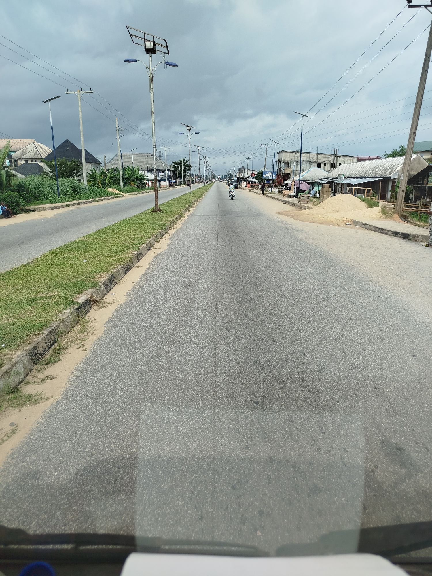 This image depicts a street scene in Yenegoa, Nigeria, on an overcast day. The road, paved with asphalt, stretches into the distance, with a median of grass and a concrete curb separating the lanes. Along the sides of the road, various buildings and structures are visible, including houses with pitched roofs, commercial buildings, and what appear to be small shops or stalls. Several utility poles with power lines are spaced along the street, and some also support solar-powered streetlights, indicating a modern infrastructure in some areas.
A few people are present, including individuals walking and a person riding a motorcycle. The overall activity is that of a typical day in a populated area, with transportation and commerce underway. The sky is cloudy, suggesting it might be morning or late afternoon, and the lighting is diffused. The presence of sand piles along the roadside hints at ongoing construction or road maintenance. The scene conveys a sense of everyday life in a Nigerian urban or semi-urban environment. There is no visible text in the image.