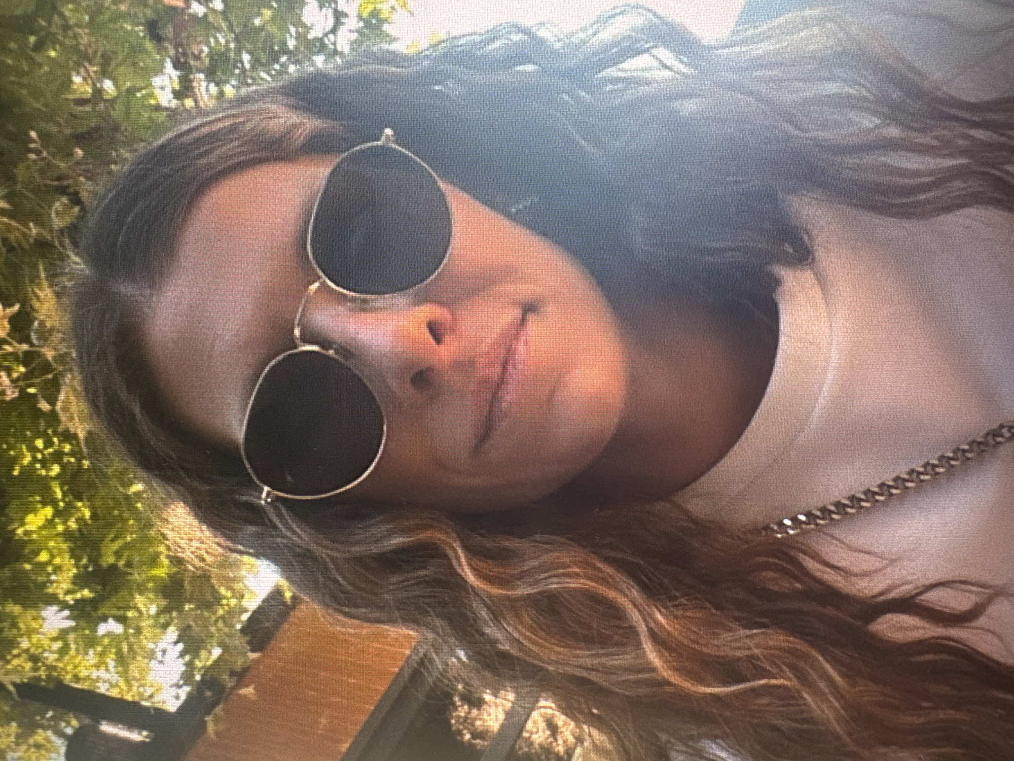 An outdoor, slightly rotated image captures a close-up of a person, likely female, in Vratsa, Bulgaria. The individual has long, wavy hair, dark at the roots with distinct lighter, possibly blonde or light brown, highlights cascading over their shoulders and chest. They are wearing round-lensed sunglasses with thin metallic frames, obscuring their eyes. A subtle, relaxed expression is visible on their lips, which are slightly parted. A simple, thin chain necklace, appearing metallic gold or silver, rests on their chest over a light-colored, possibly off-white or cream, T-shirt or top.
The background is filled with lush green foliage and tree leaves, suggesting a sunny daytime setting in a natural environment such as a park or garden. Hints of a wooden structure or furniture are also visible in the lower part of the frame. The lighting indicates a bright, sunny day. The image appears to be a selfie taken from a slightly upward and tilted angle, and it exhibits some pixelation or digital screen-capture artifacts, particularly noticeable in the detailed crops. There is no visible text in the image.