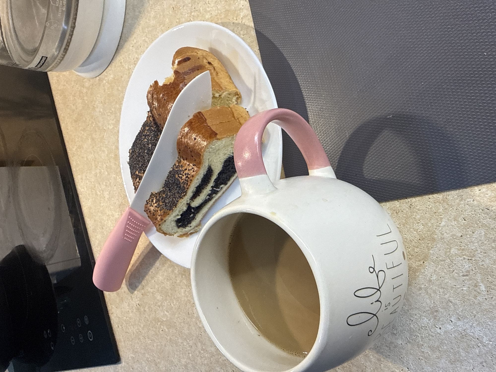 A top-down view shows a scene featuring food and drink. A white plate holds slices of poppy seed loaf and a pink-handled knife rests atop the bread. A white mug with a pink handle contains a tan liquid, likely coffee or tea. The background consists of a light brown countertop, a black cooktop, and a dark gray surface. The location appears to be a kitchen setting of Unknown, Unknown.