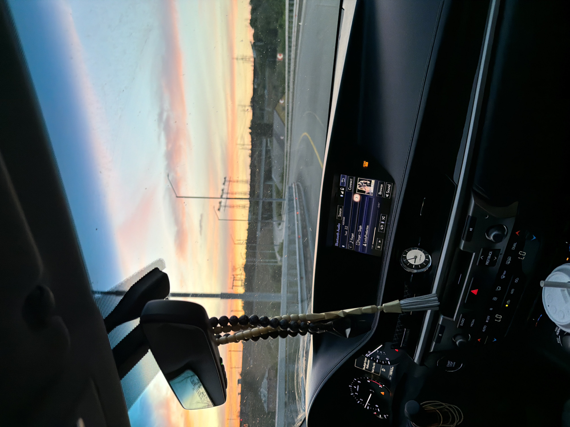 This media file captures an interior view of a car, likely a Lexus, during a sunset in Zaporizhzhia, Ukraine. The sun is setting, casting a vibrant palette of blues, pinks, and oranges across the sky, visible through the windshield. The car's dashboard is prominently featured, displaying the infotainment system with the text "Bluetooth Audio," "Track 22," and song titles like "Forget" and "Lucky Production." A small analog clock with Roman numerals is also visible on the dashboard, indicating a time that appears to be around 3:15. Prayer beads hang from the rearview mirror, suggesting a personal or spiritual element to the journey. The driver's side dashboard includes various controls and a speedometer, showing a mileage of 150762. The overall mood is serene, with the beautiful sunset offering a moment of calm during what appears to be a drive. The weather seems clear, conducive to enjoying the scenic view.
