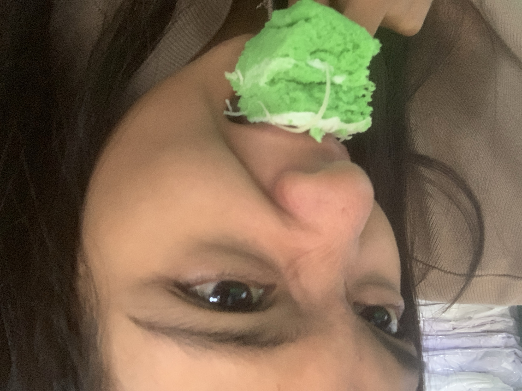 A close-up, eye-level, medium shot shows a person about to bite into a bright green piece of cake with white frosting. The person's face is partially obscured by the cake. The cake is held by the person's right hand. The person has dark hair, dark eyebrows, and dark eyes. The person is wearing a light brown shirt. The background is blurry but appears to be an indoor environment with stacks of white items. There are no visible indications of the location in the city of Sungai Meriam, Indonesia.