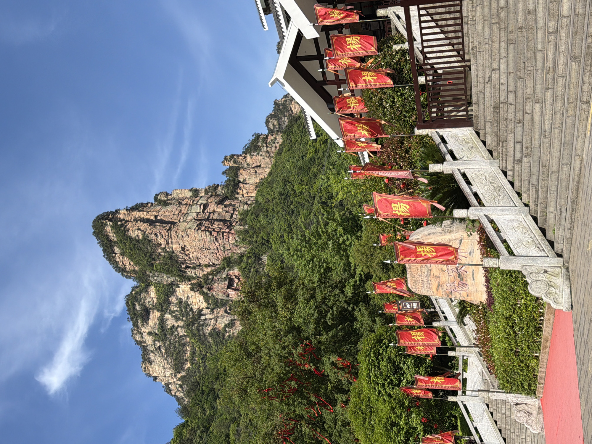 The image shows a mountain landscape with a stone staircase lined with red flags bearing gold characters. The staircase leads up to a building with a white roof. The mountain is covered in green trees and shrubs, and the sky is blue with a few clouds. The location is likely in the city of Zhonghu, China.