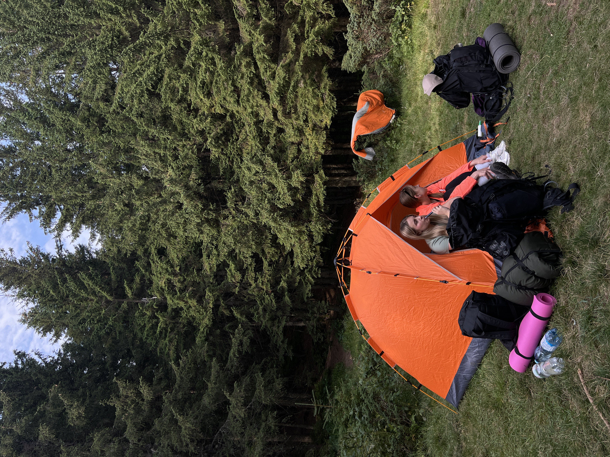 In a natural environment resembling a forest clearing, two young women are seated inside an orange tent, suggesting a camping or outdoor recreational activity. The tent is set up on a grassy area surrounded by tall evergreen trees. Outside the tent, several pieces of camping equipment are visible, including backpacks, sleeping bags, a pink sleeping mat, and water bottles, indicating a prepared expedition. A folded sleeping bag is also seen hanging on a bush in the background. The overall scene portrays a tranquil outdoor setting, possibly in or near the city of Vorokhta, Ukraine, a region known for its natural beauty and forested landscapes.