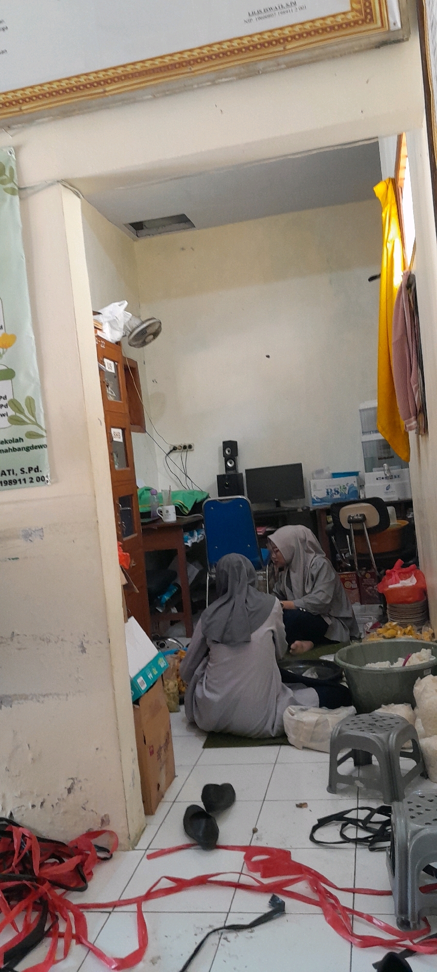 The image depicts an indoor scene, likely a room in Lemahbang Dewo, Indonesia. Two individuals are seated on the floor, facing each other. Both are wearing hijabs. The individual on the left is wearing a grey tunic and head covering, while the person on the right is wearing a grey jacket and head covering. They appear to be interacting.
The room has tiled flooring with a white and beige color scheme. There are various objects scattered around. To the left, a partially visible banner with green foliage and text hangs on a cream-colored wall. Near the banner, a wooden cabinet with multiple compartments is visible. A ceiling fan is mounted on the wall above the cabinet.
In the central area of the room, there is a wooden table with items on it, including a mug, a green cloth, and a bottle. Behind the table, a blue chair and a computer monitor are present. Speakers are stacked next to the monitor. To the right, plastic storage containers are stacked, and a yellow fabric hangs on the wall.
The foreground of the image shows scattered red and black plastic ribbons, and a pair of black slippers. Several grey plastic stools are also visible. A large green plastic tub containing white material is situated to the right of the individuals. The walls show signs of wear and tear, with peeling paint in some areas. The overall environment suggests a functional space that might be used for storage or communal activities.