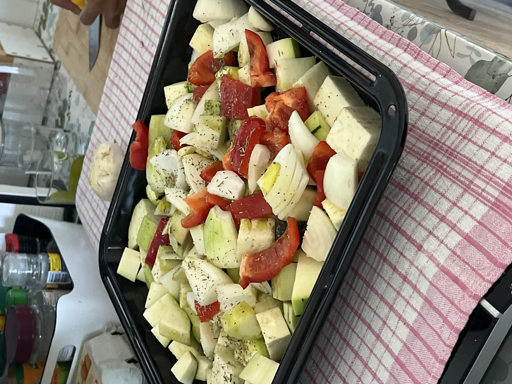 A close-up shot shows a black baking tray filled with chopped vegetables, including zucchini, red bell peppers, and onions, seasoned with herbs. The tray is placed on a pink and white checkered cloth, which in turn rests on a patterned tablecloth. In the background, out of focus, are jars of spices and a glass of water, suggesting a kitchen environment. The scene is indoors. The city of Montélimar, France is not visible in the image.