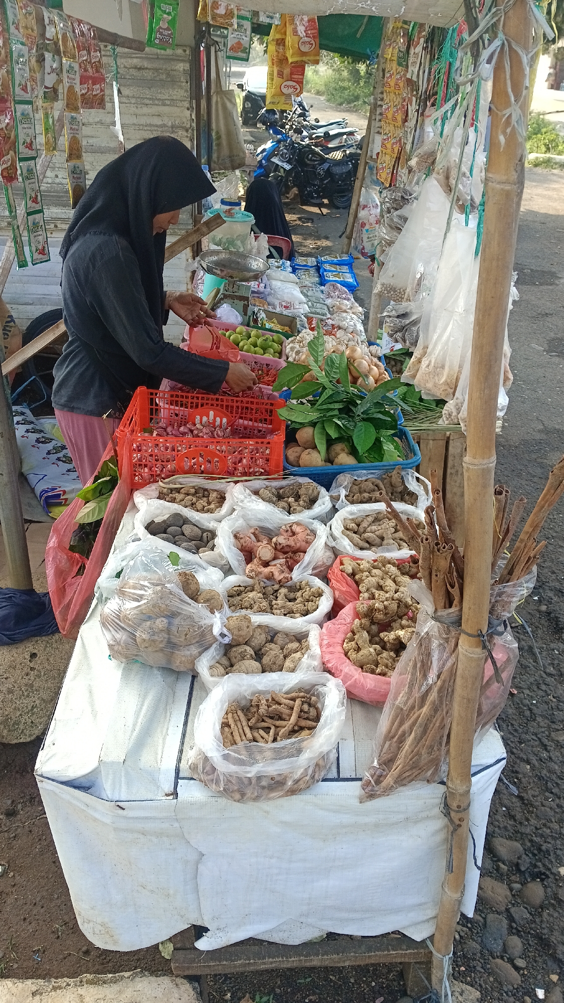 A street-side vendor stall in Tajurhalang, Indonesia, displays a variety of fresh produce and dried goods. A woman wearing a black hijab and a dark grey top is seen arranging items in a red plastic crate filled with garlic. The stall is covered by a green tarp, and various packaged goods hang from the structure. Below the tarp, the stall features a white cloth-covered table laden with an array of items. These include bags of what appear to be medicinal roots and tubers, such as galangal, ginger, and turmeric, along with cinnamon sticks bundled together. Other produce, like limes and onions, are visible in plastic crates. In the background, motorcycles are parked, and a car is partially visible, suggesting the stall is located along a road. The ground surrounding the stall is unpaved, with visible stones and dirt.