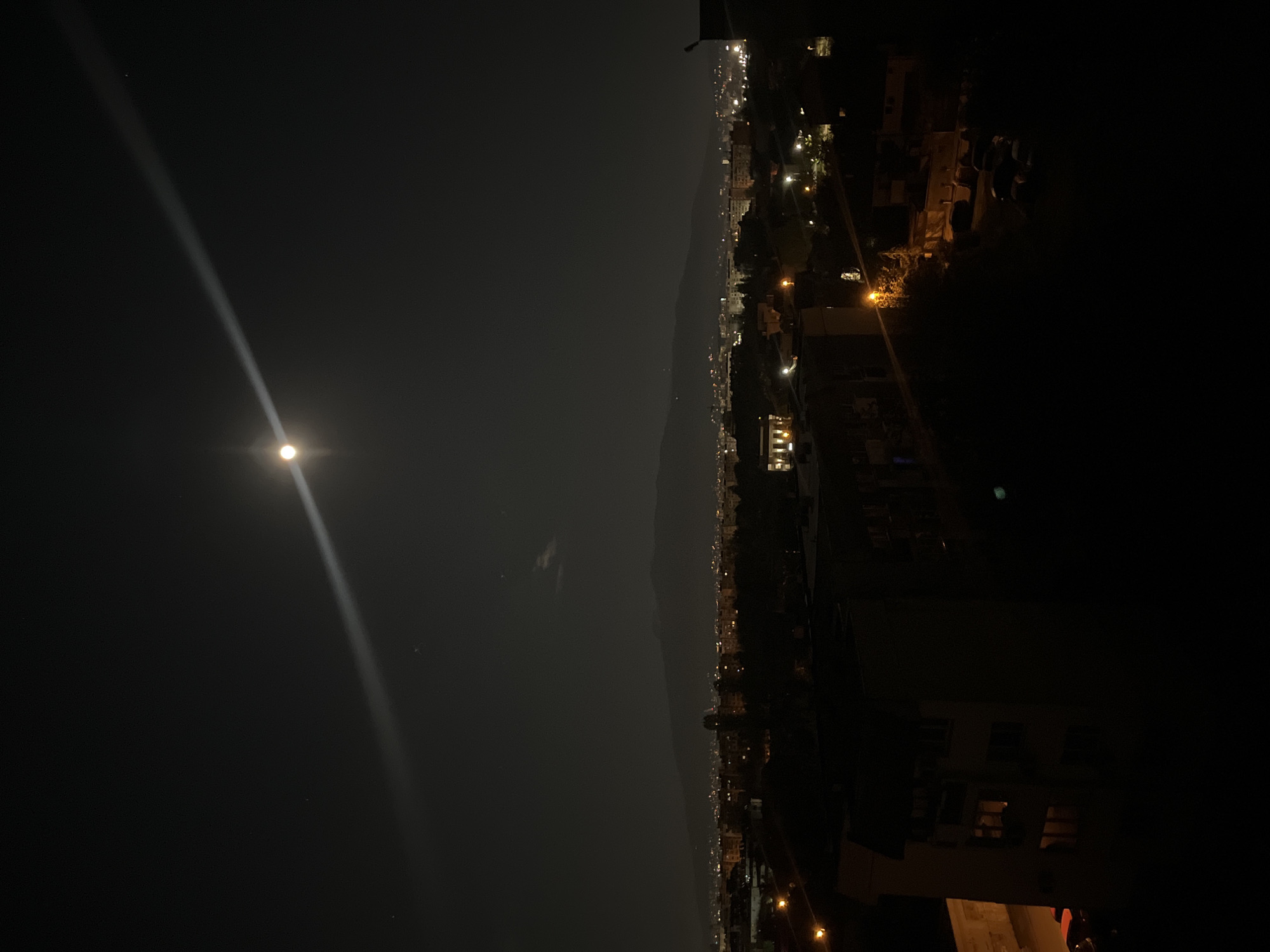The image depicts a nighttime cityscape. The moon is prominently visible in the sky, with a light beam extending upwards. A mountain range or elevated terrain runs horizontally across the middle of the frame. Below this, a city with scattered lights is partially visible. The foreground is dark with some structures, possibly buildings, barely discernible. There is no readily identifiable information in the photo to pinpoint this scene to Sofia, Bulgaria.