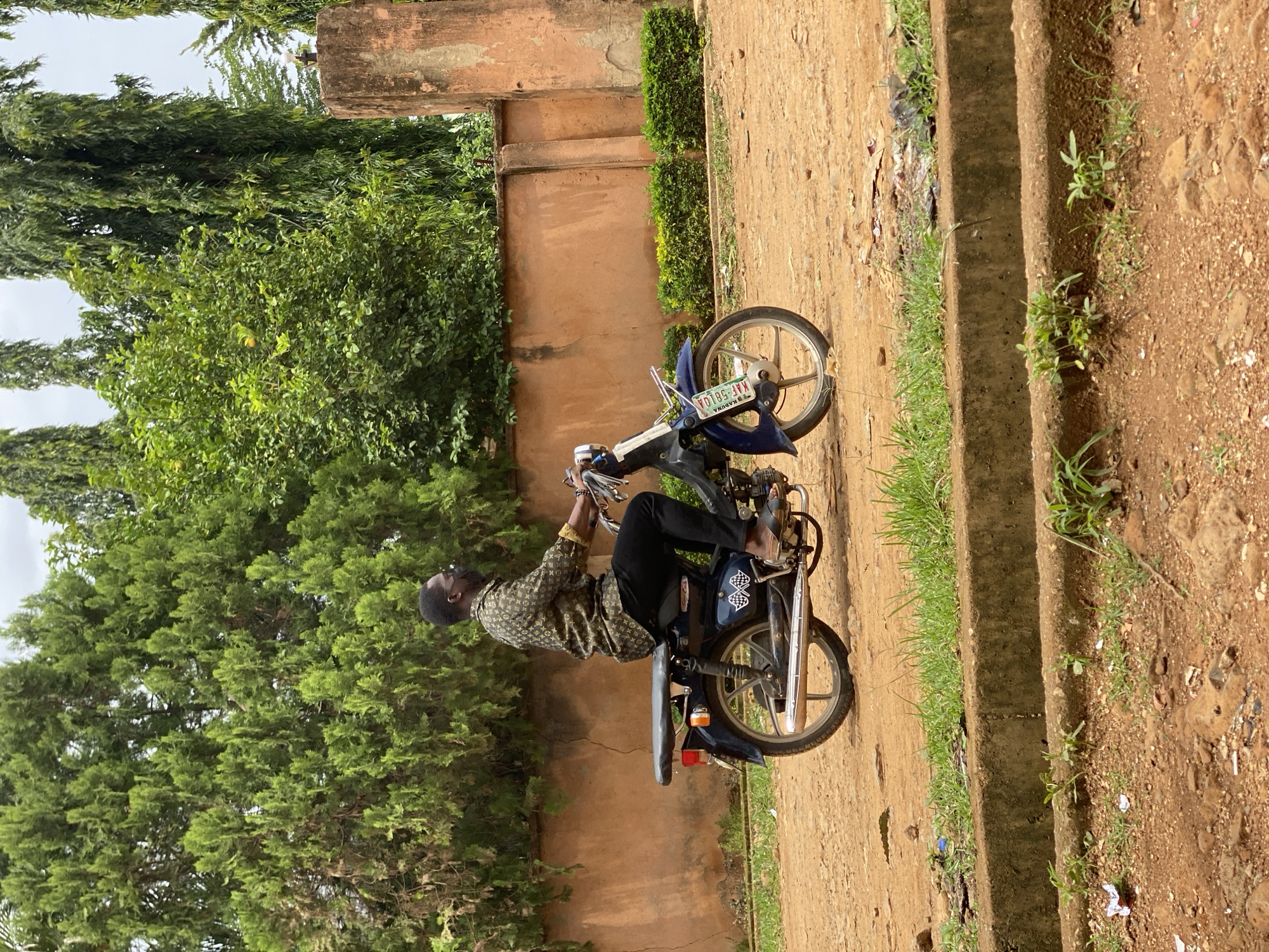 A man is sitting on a blue motorcycle in what appears to be an outdoor setting in Maigizo, Nigeria. The environment includes vegetation with various trees, a building with a brown wall, and an unpaved ground area. The motorcycle has a license plate. The man is wearing black pants and a patterned shirt, with sandals on his feet.