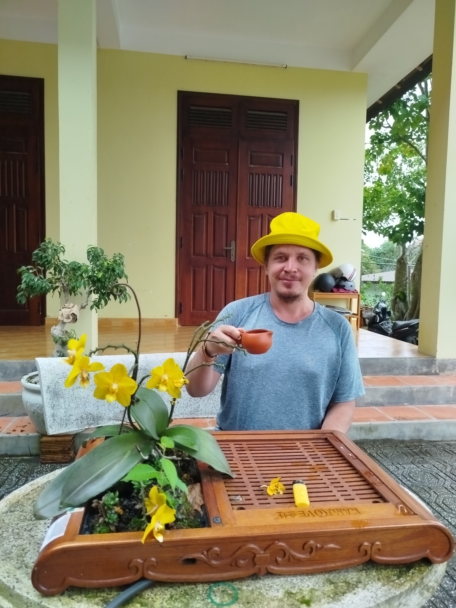 A Caucasian man wearing a yellow bucket hat and a grey t-shirt holds a small, brown ceramic tea cup in his right hand. He is seated at a wooden table with a tea tray. A potted orchid with yellow flowers and a small bonsai tree are also on the table. The man is smiling and looking at the camera. In the background, there are two wooden doors and a covered patio area. The scene appears to be outdoors, likely in a residential setting in Da Lat, Vietnam, given the architecture and lush greenery. The lighting suggests it is daytime, possibly overcast.