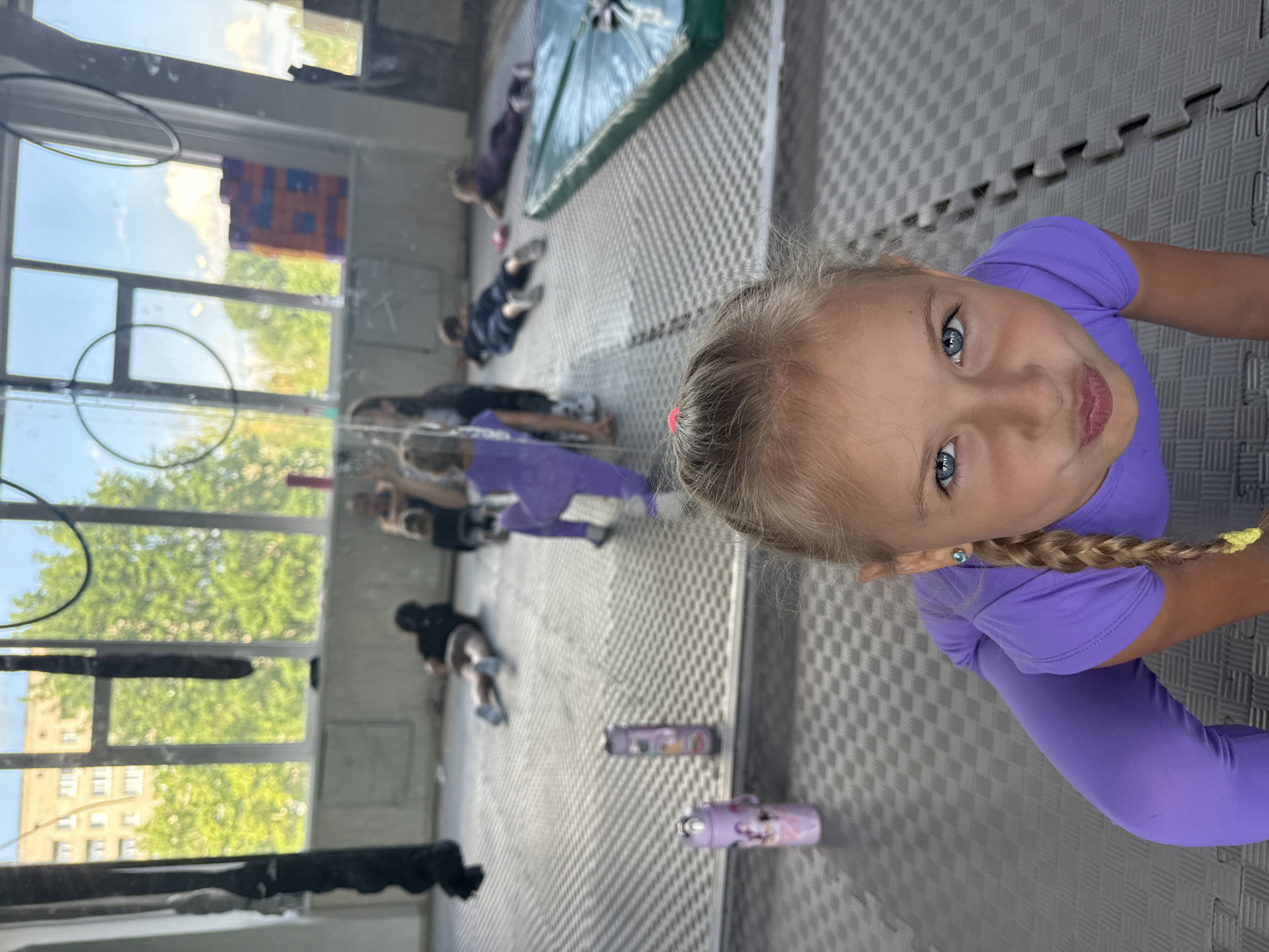 The image shows a young girl with blonde braided hair and blue eyes, wearing a purple leotard. She is looking up at the camera with a playful expression. The setting appears to be an indoor gym or studio, with padded grey floor mats and large windows reflecting the outside. Several other children and adults are visible in the background, lying on the mats, possibly participating in a gymnastics or exercise class. The windows offer a view of trees and a building, suggesting it's daytime and likely clear weather. There are also some gym equipment visible, including what looks like a hoop and a mat covered in green material. A few purple water bottles are scattered on the floor. The overall mood of the image is lighthearted and active.