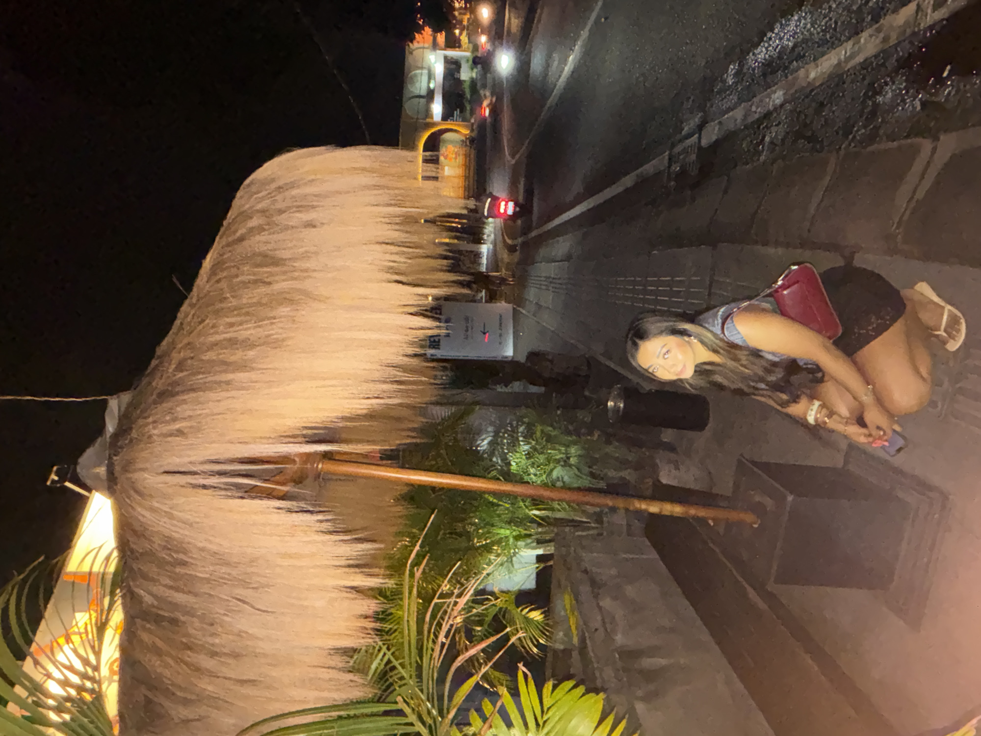 The image depicts a nighttime street scene in North Kuta, Indonesia. A woman is crouched on a sidewalk next to a large thatched umbrella, looking towards the camera. She is wearing a dark lace skirt and a light-colored top, with a red handbag slung over her shoulder. In the background, a scooter is seen driving on the street, along with other vehicles and illuminated buildings. Palm trees and lush vegetation are visible to the left of the scene. The overall ambiance suggests a lively evening in a tropical location.