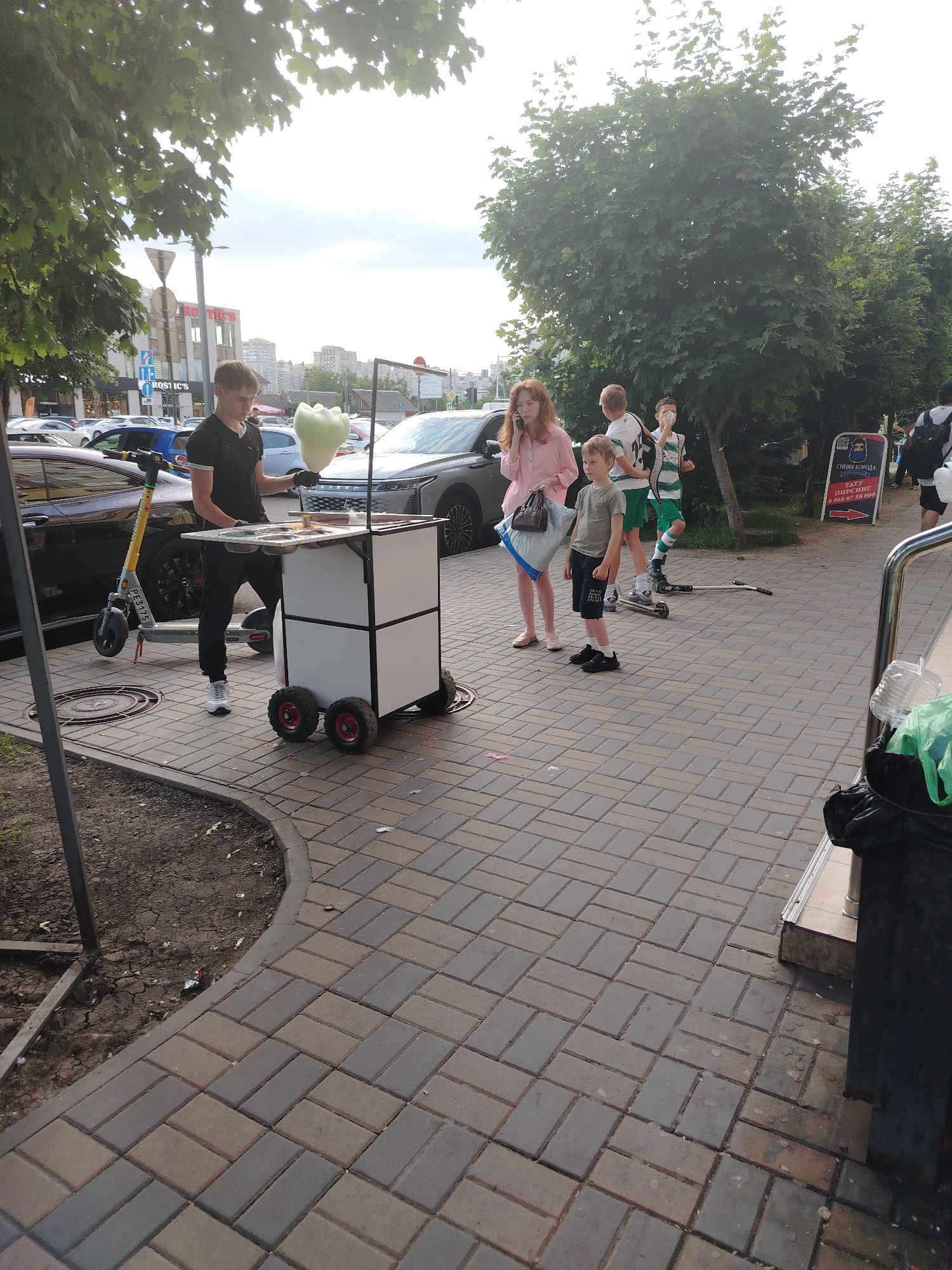 A young male with short brown hair, wearing a black t-shirt and black pants, stands behind a white cart with wheels. The cart has a white top surface with three containers. The male is holding a pale green, fluffy object that resembles cotton candy. To his left, a grey electric scooter with yellow accents is parked. In the background, there are several cars parked on the street, including a dark-colored sedan and a silver SUV. A woman in a pink shirt and a young boy in a green shirt and dark shorts stand to the right of the cart, looking in its direction. Two other boys are visible behind them, one wearing a green and white striped shirt and the other a white shirt. The scene appears to be on a paved sidewalk in a city. The buildings and trees in the background suggest an urban environment. The presence of the cotton candy cart and the electric scooter indicates a public area, possibly a park or a commercial street.