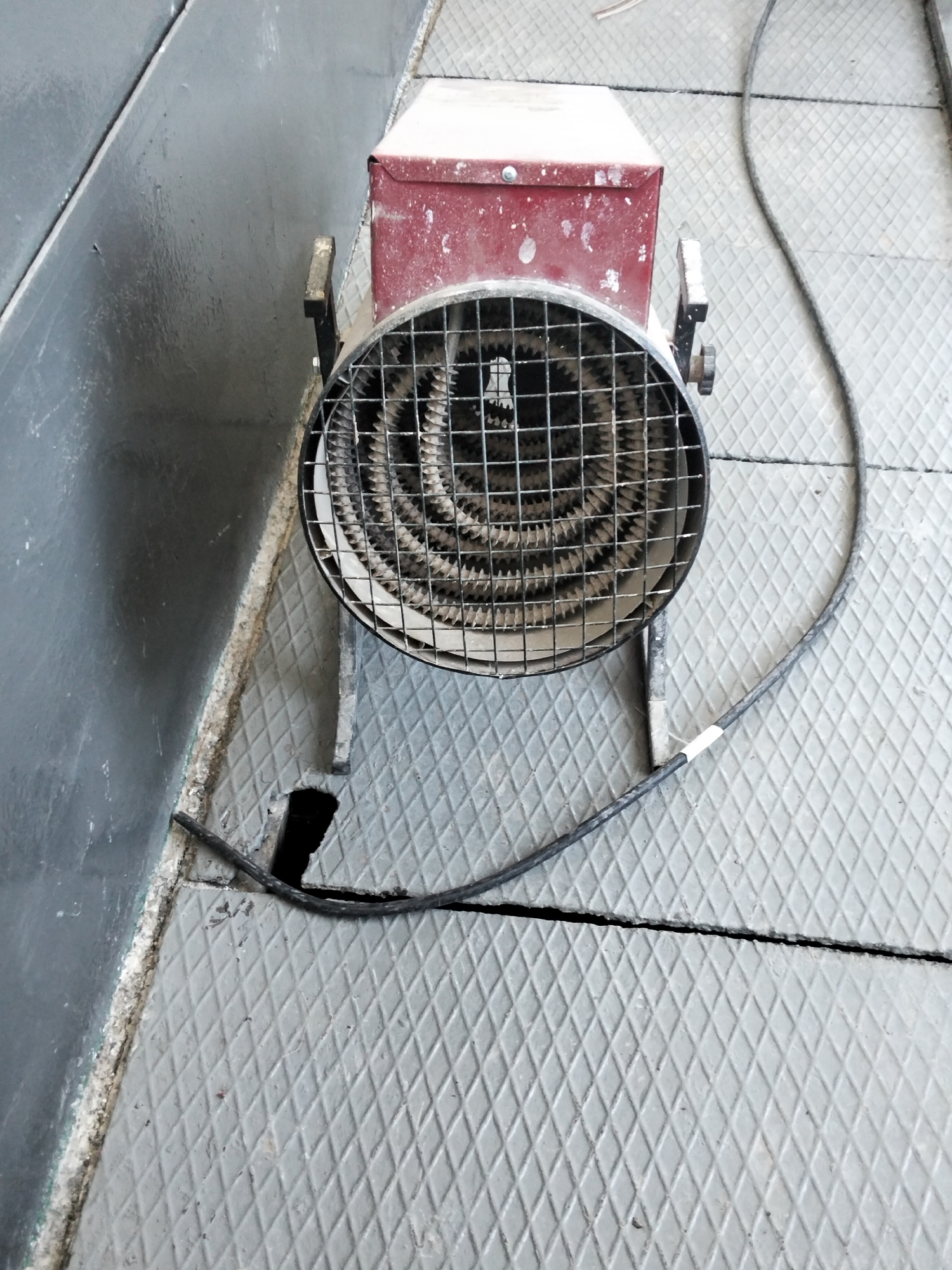 An electric heater with a red metal housing and a wire grid protecting the heating element is placed on a diamond-patterned metal floor. A black power cord extends from the heater and lies on the floor. The heater is positioned next to a dark gray wall. The background shows a continuation of the metal floor. There are no people visible in the image. The scene appears to be indoors, possibly in an industrial or workshop setting. The location is identified as Батыр ауылдық округі, Kazakhstan.
