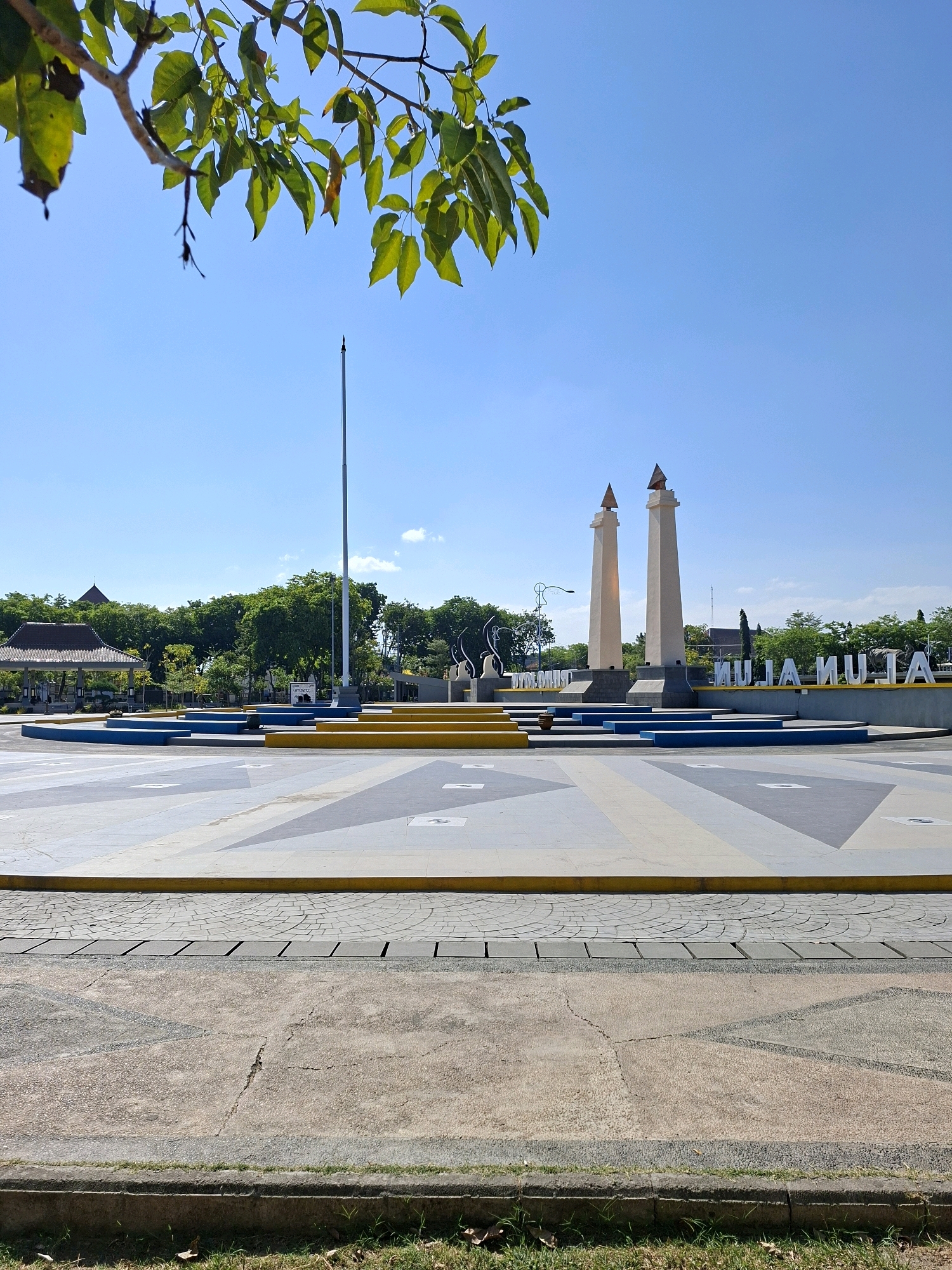 The image captures a public square in Gunung Sekar, Indonesia, under a clear blue sky. In the foreground, the ground is paved with a patterned concrete surface, separated by a concrete curb and some sparse grass. Moving further into the scene, there is a raised, multi-tiered platform with yellow and blue accents, leading towards a central monument. The monument features two tall, slender, cream-colored pillars topped with triangular elements. Behind the pillars, large white lettering spells out "ALUN ALUN". In the background, lush green trees form a backdrop to the monument, and to the left, a traditional Indonesian pavilion with a tiled roof can be seen. A flagpole stands tall in the center of the square, to the left of the monument. The overall impression is of a well-maintained, open public space.