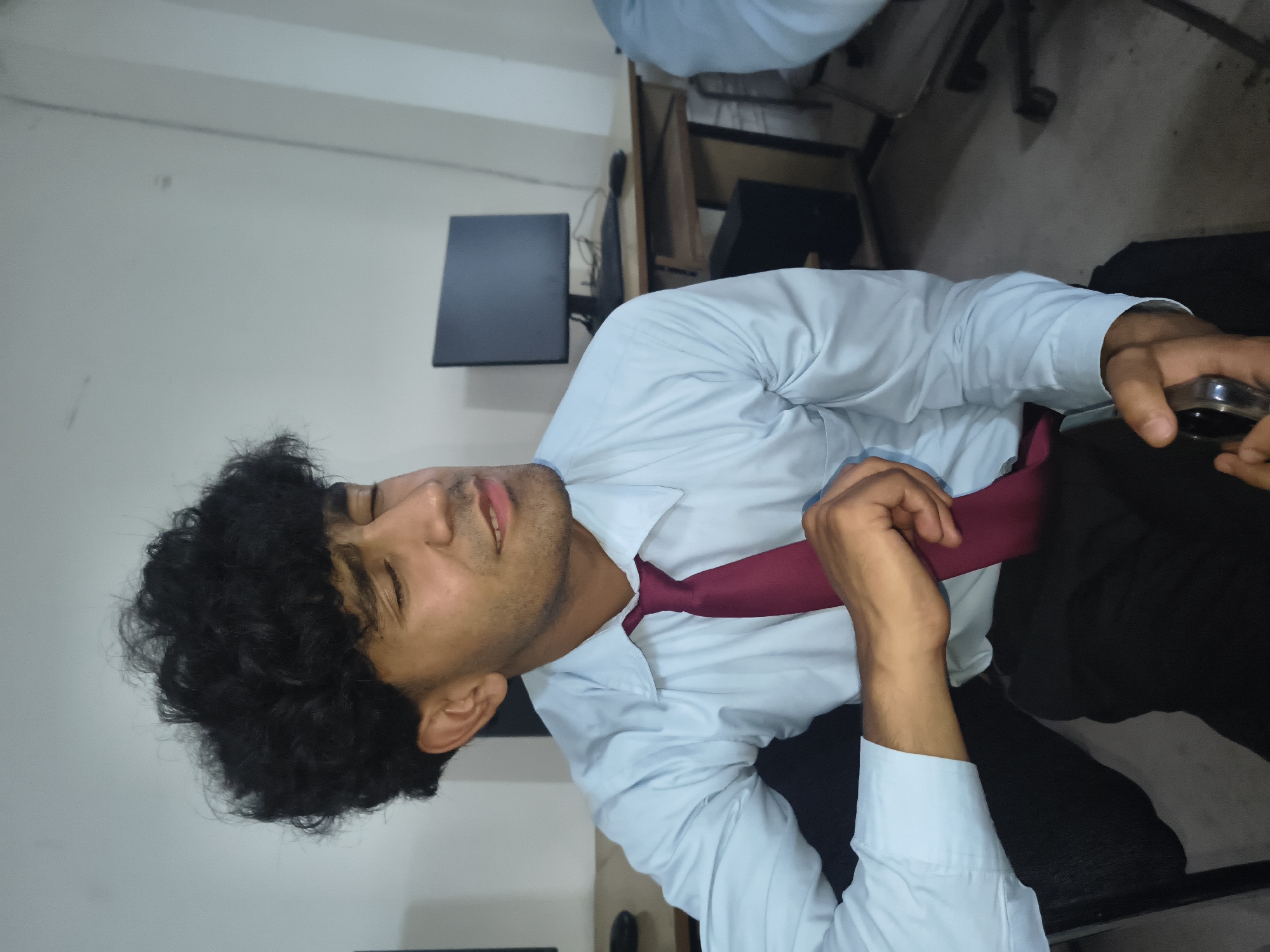 A young man with dark, curly hair is seated in a room with light-colored walls and a tiled floor. He is wearing a light blue collared shirt and a dark red tie. His eyes are closed, and his head is tilted back slightly, suggesting he might be listening to something or in thought. His right hand rests on his tie, while his left hand is holding a silver object, possibly a phone. In the background, there are computer monitors and desks, indicating the room could be an office or a computer lab. The overall lighting is dim, with a slight yellow hue. The man's expression is relaxed. This scene is not specific to Dharamshala, India, and could be found in any similar indoor setting.