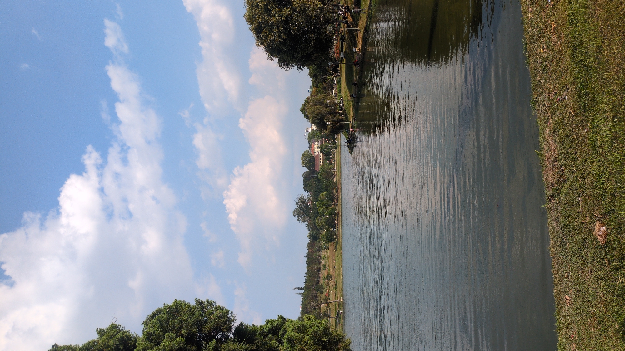 The image depicts a view of Xuan Huong Lake in Da Lat, Vietnam. The lake occupies the central portion of the image, reflecting the sky and surrounding scenery. Along the left side of the lake is a grassy bank and trees. On the right side, there is a bank of grass leading to a line of trees. Buildings can be seen in the distance beyond the trees. The sky above is light blue with scattered white clouds.