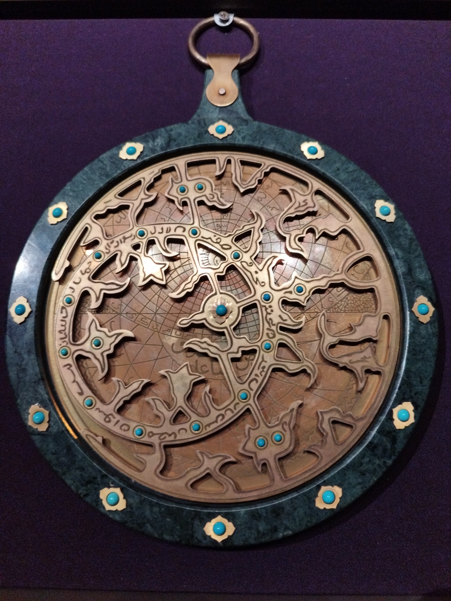 This is a close-up, eye-level, full-frame shot of an astrolabe, a historical astronomical instrument. The astrolabe is circular, with a dark green, marbled outer rim. The rim is adorned with small, gold-colored, flower-shaped embellishments, each containing a bright turquoise gem at its center. The central part of the astrolabe is made of a copper-colored metal and features intricate geometric patterns and what appear to be celestial charts or constellations. There are many small turquoise gems embedded within this central design, highlighting various points. A metallic ring, likely for suspension, is visible at the top of the instrument. The background is a deep purple fabric, and a dark border frames the entire image. The lighting is somewhat dim, casting soft shadows and giving the astrolabe a museum-quality presentation. The location is Unknown, Unknown.