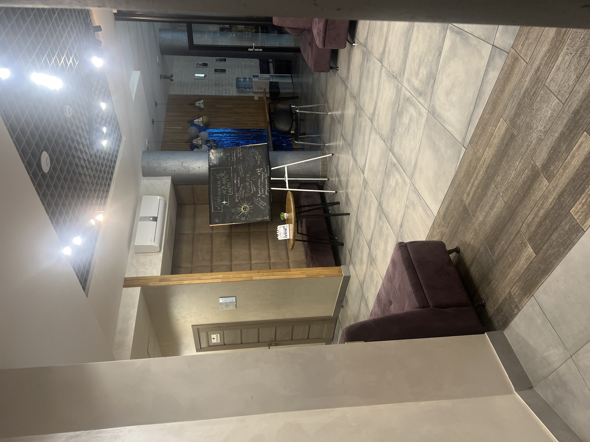 The image depicts an indoor scene, likely a reception area or lounge, in what appears to be a modern establishment. The environment is characterized by a tiled floor transitioning from large, light gray square tiles to wood-look planks. The walls are painted in neutral tones, with one wall featuring a textured padded brown finish. A dark purple upholstered bench seat is visible in the foreground.
Further into the space, a dark wooden door with a "RESTROOM" sign is positioned to the left. In the center, a blackboard on an easel displays handwritten messages and drawings, suggesting it's a point of interaction or information sharing. Balloons and shimmering blue tinsel are draped behind the blackboard. Several black wire-frame chairs with gray seats are arranged around a table and near a water cooler. The ceiling is a suspended grid with recessed lighting.
The overall impression is one of a clean, contemporary space, possibly a hotel lobby, office waiting area, or similar commercial establishment. The presence of the "RESTROOM" sign strongly suggests it is a public or semi-public indoor area. The scene is devoid of any visible people, focusing solely on the architectural elements and furnishings. The lighting is bright and evenly distributed, indicating artificial illumination is the primary source.